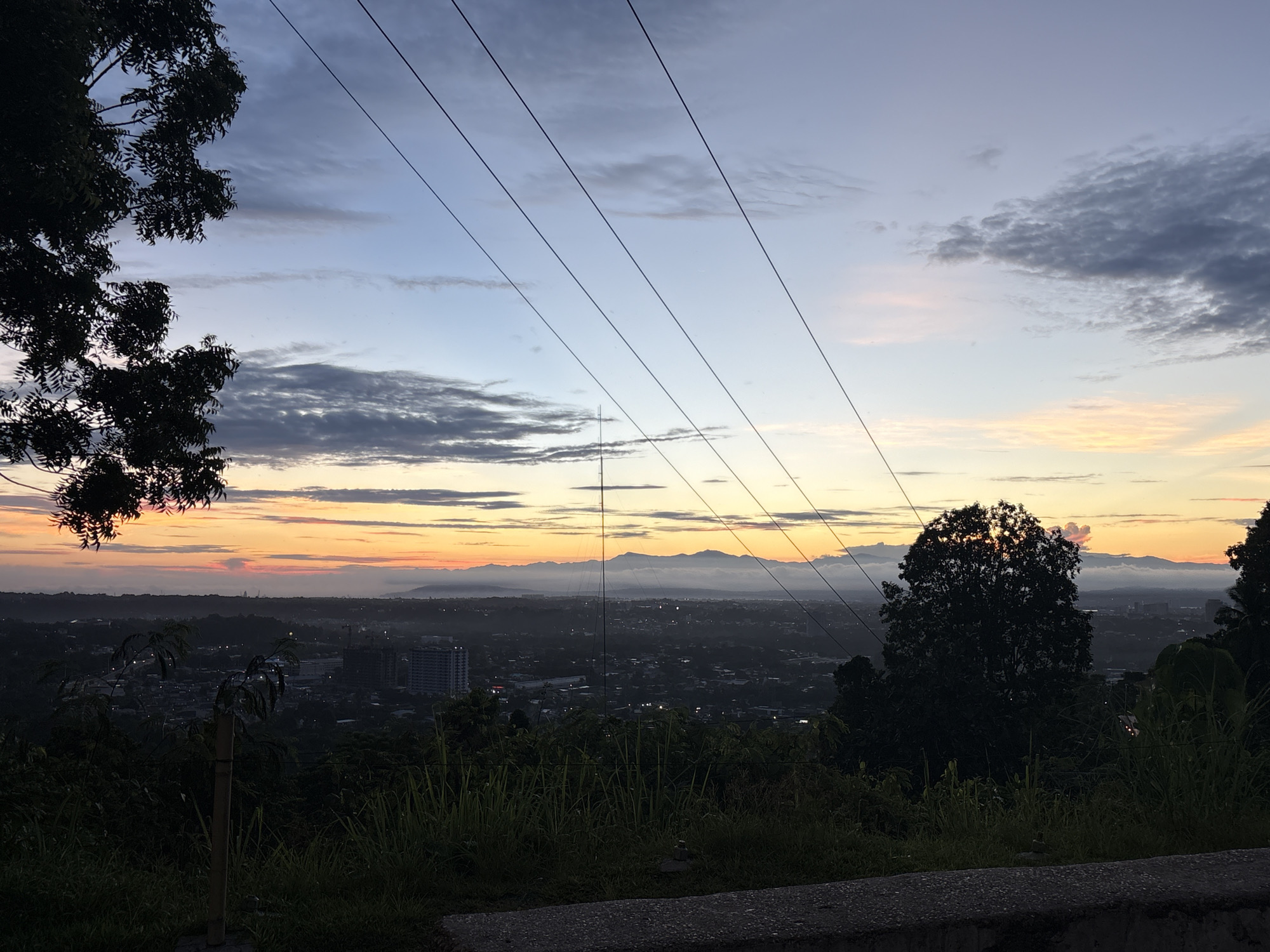 The image captures a panoramic view of Davao City, Philippines, at dusk. In the foreground, the silhouette of lush green vegetation and a few trees frames the scene, with a low concrete barrier visible at the very bottom. A few utility poles and wires stretch diagonally across the sky, breaking the horizon. The midground reveals the sprawling cityscape, with buildings and houses dimly lit against the darkening landscape. In the far distance, a range of mountains is visible, their peaks softened by the twilight haze. The sky is a gradient of colors, transitioning from a soft orange and pink near the horizon to a deep blue overhead, with scattered clouds adding texture. The overall atmosphere is serene and tranquil, characteristic of a sunset in a tropical city.