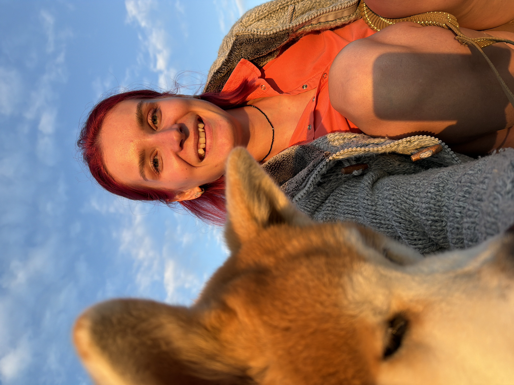 A young adult female with bright red hair and light skin, illuminated by warm, directional sunlight, is positioned on the right side of the frame. She is smiling broadly, exposing her teeth, and looking directly towards the camera. Her left ear displays multiple piercings with small gold hoop earrings. She wears a black beaded choker necklace, an orange collared shirt, and a grey knitted cardigan with visible wooden toggle buttons. Her right knee is bent and prominent in the lower right, casting a distinct rectangular shadow, likely from a handheld device. A gold-colored, possibly patterned fabric garment is visible on her right leg below the knee, with a thin rope-like tie.
In the blurred foreground, occupying the left and central portions of the image, is the head and upper body of a light brown and cream-colored dog, likely a Shiba Inu, highly out of focus due to shallow depth of field. Its pointed ears and fur are distinctly visible, with strong golden highlights from the sunlight. The dog is positioned very close to the woman, partially obscuring her left side. The background consists of a clear blue sky with scattered white cumulus clouds. The lighting indicates a late afternoon or early evening setting, characterized by intense golden hour illumination. The image provides no visual information to confirm a specific location within the city of Chaikovsky, Russia, or any other geographical area.