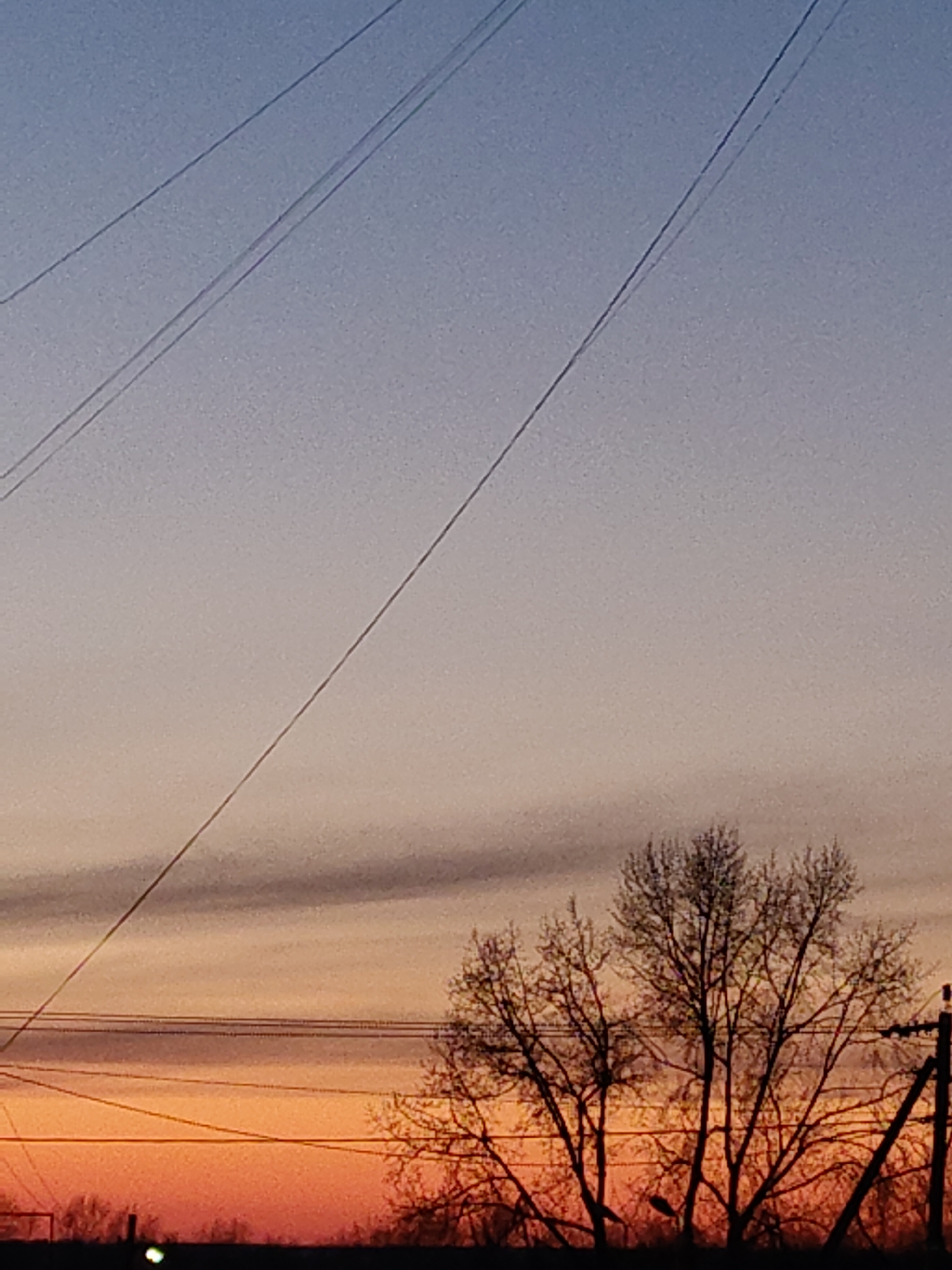 The image captures a twilight or sunset scene. The sky transitions from a gradient of blue at the top to orange at the bottom. Bare branches of a tree are silhouetted against the colorful sky. Utility poles and wires are visible, adding to the urban or suburban feel. The location is identified as Unknown, Unknown.