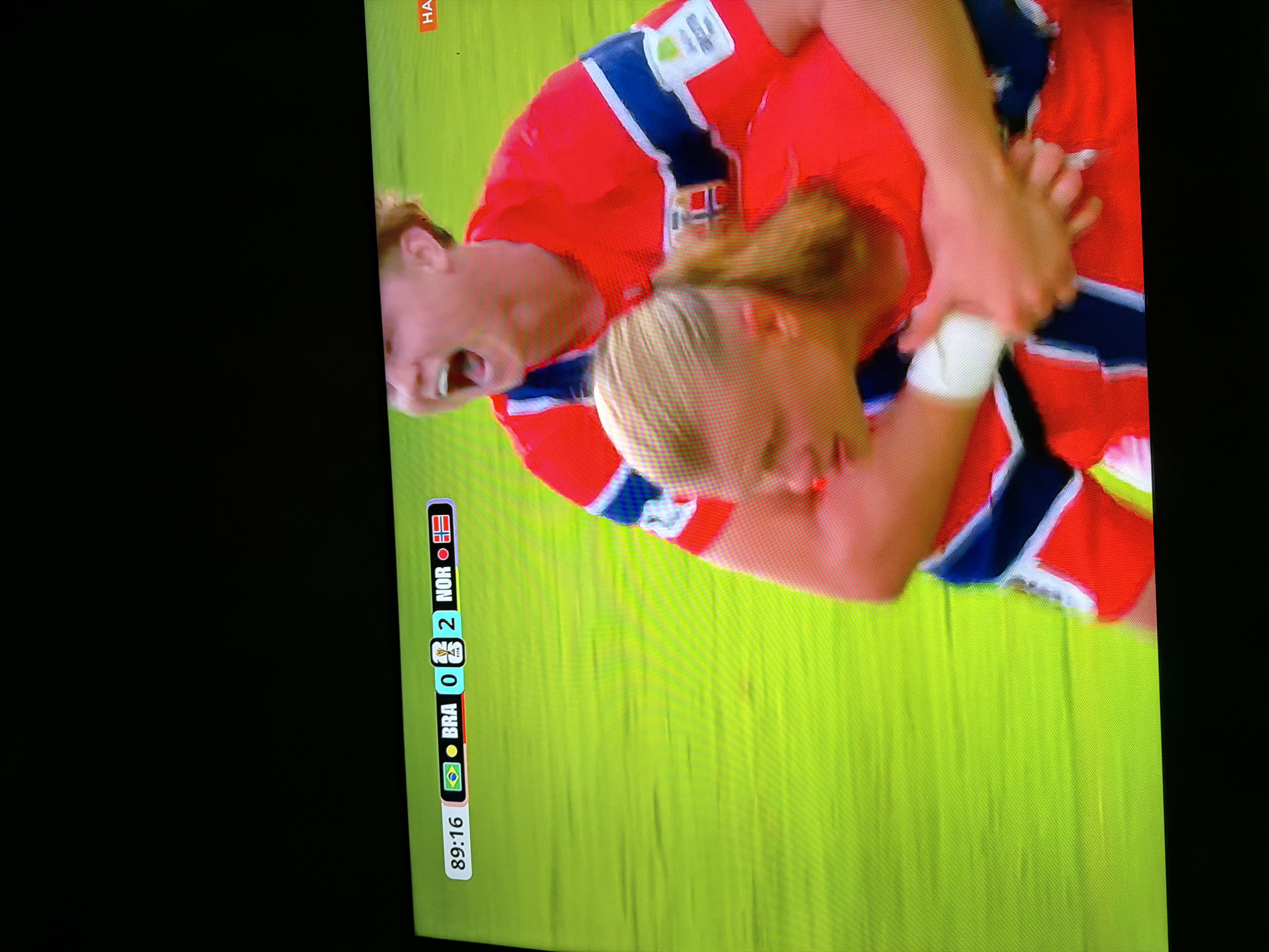 An overhead shot shows a soccer match with a score of Brazil 0, Norway 2. The time is 89:16. Two players in red and blue uniforms are celebrating. One player is on the shoulders of another, with both players smiling and cheering. The background is a bright green field. The image appears to be taken from a television screen. There is no information in the image to suggest this is in Ognianovo, Bulgaria.