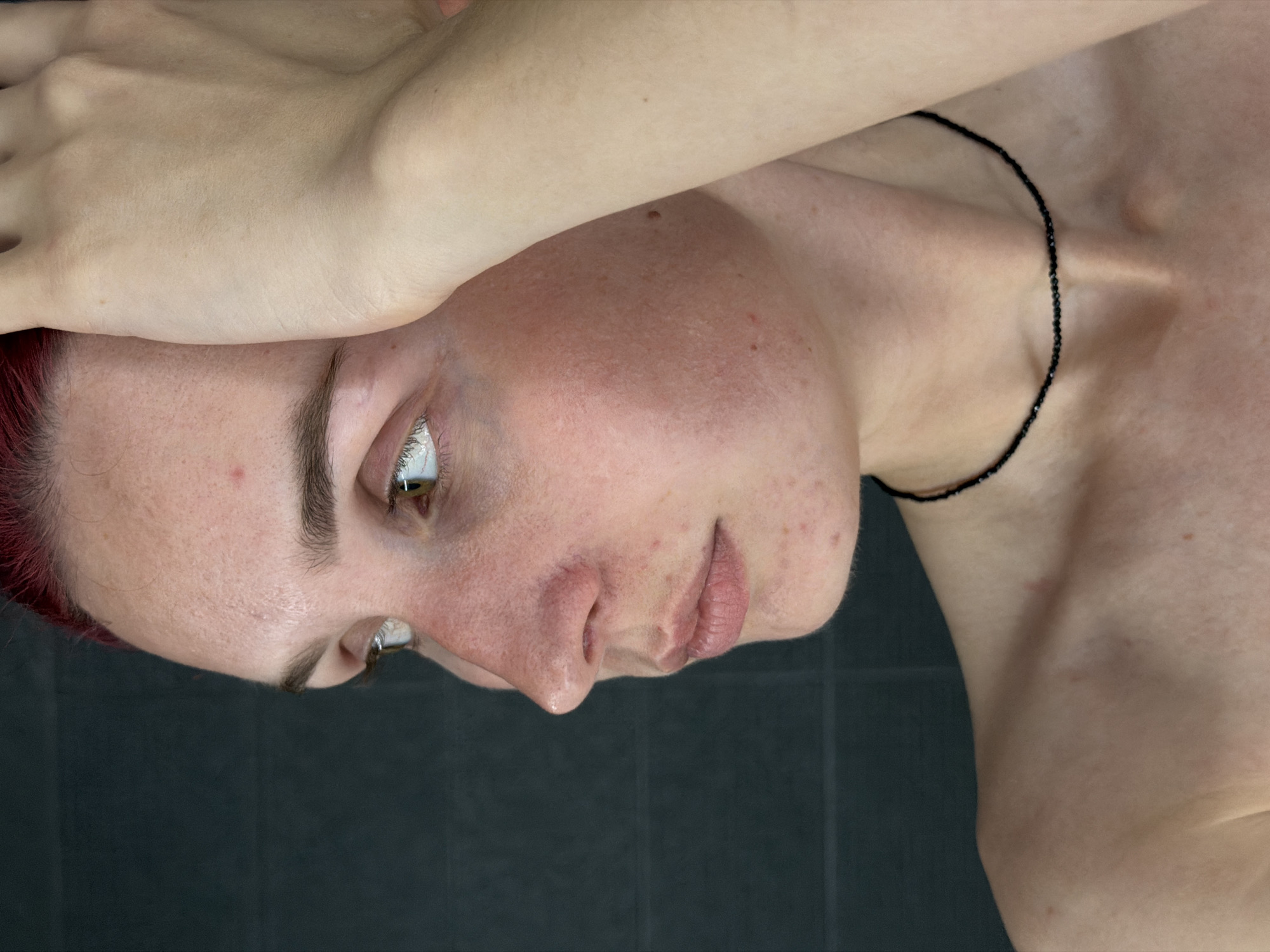 A profile close-up shows a young person with fair skin and red hair. They are wearing a black beaded necklace. The person is holding their right hand to their forehead. The background is a dark, textured wall. The scene does not provide any information about the location in Chaikovsky, Russia.
