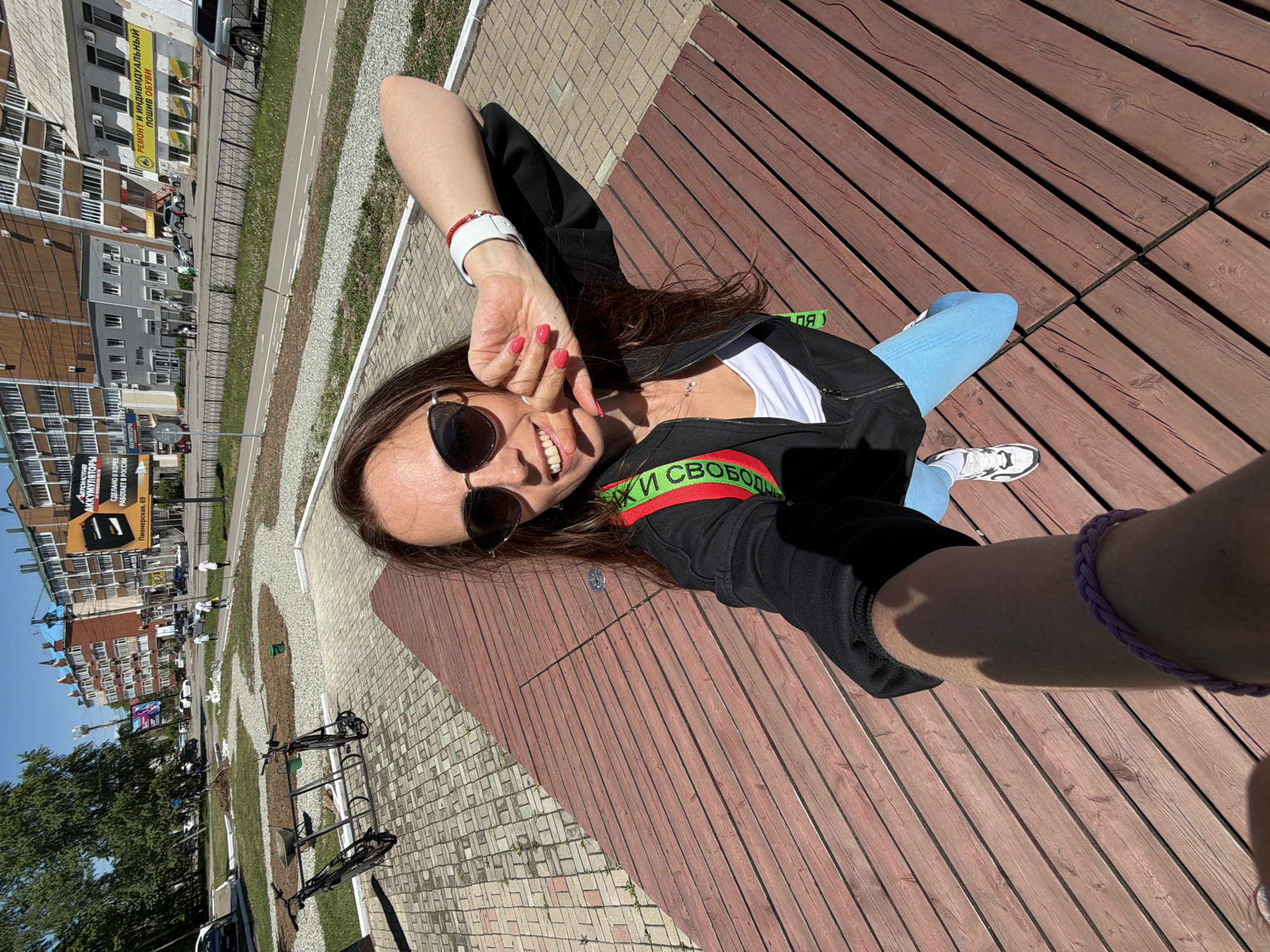 A young woman is prominently featured in an outdoor selfie, smiling broadly at the camera. She has long dark hair and wears sunglasses, a white top, a black zip-up jacket with a green and red stripe, and light blue leggings paired with white sneakers. Notable accessories include red nail polish, a white wristband, a red beaded bracelet, and a purple braided bracelet. The text "К СВОБОДЕ" (To Freedom) is visible on the green and red stripe of her jacket.
She is situated on a wooden deck or large wooden planks, possibly part of a public seating area, adjacent to a paved path made of cobblestones or bricks. The scene unfolds under a clear blue sky with bright sunlight, casting distinct shadows, indicating daytime. The background shows an urban environment with a street, several cars, and multi-story buildings. Greenery, including trees and grass, and a bicycle rack with bicycles are also visible.
In the background, on various buildings, several signs with Cyrillic text are present. One yellow banner advertises "РЕМОНТ И ИНДИВИДУАЛЬНЫЙ ПОШИВ ОБУВИ" (Repair and Individual Tailoring of Shoes). Another billboard promotes "АККУМУЛЯТОРЫ" (Batteries), stating "СДЕЛАНО В КОРЕЕ РАБОТАЕТ В РОССИИ" (Made in Korea, Works in Russia) and displaying "Пионерская, 69" (Pionerskaya, 69) as an address. The woman appears relaxed and happy, enjoying the sunny weather in this city setting in Birobidzhan, Russia.