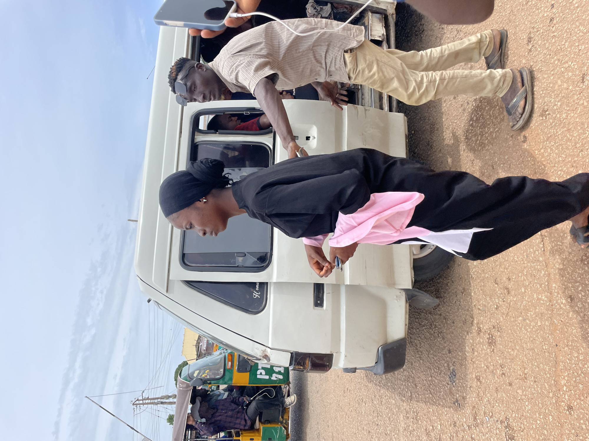 In Gura Top, Nigeria, an outdoor scene unfolds under a cloudy sky. A woman, draped in a black abaya and a black headscarf, stands to the left of a white van. A pink and white fabric is draped over her left arm. To her right, a man wearing a striped shirt and light tan pants leans towards the open side door of the van, his right arm extended. He wears dark sunglasses. Inside the van, another person wearing a red shirt is visible. In the background, a green and yellow auto-rickshaw is parked next to the van. The ground is covered in reddish-brown gravel. Electrical poles and wires are present in the distance, suggesting an urban or semi-urban environment.