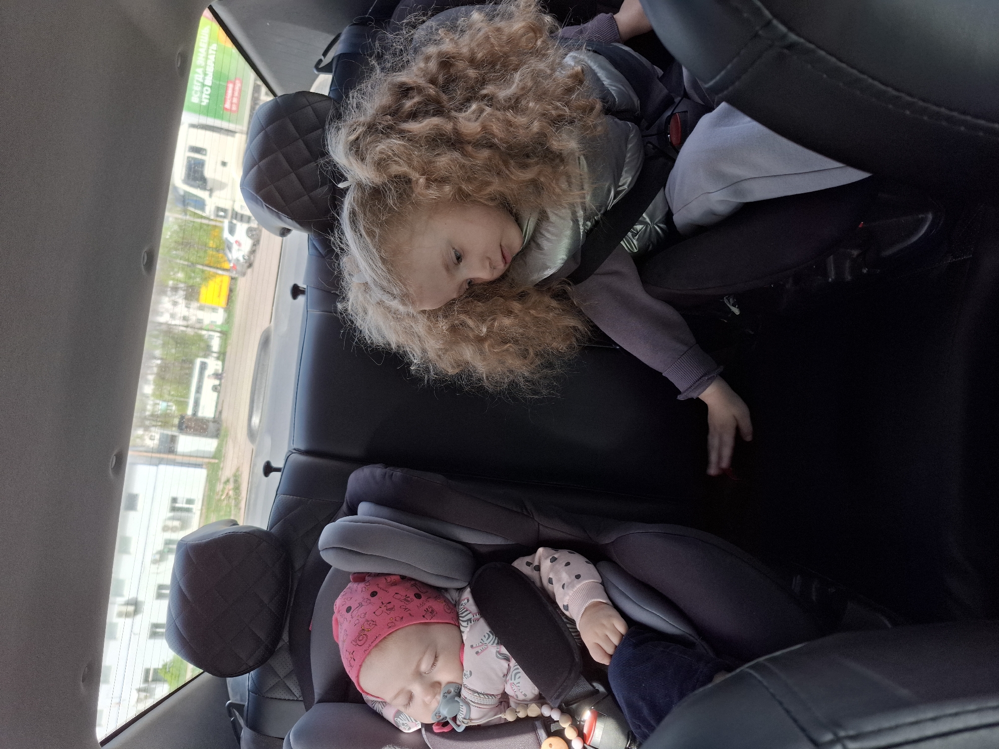In the back seat of a vehicle in Chaikovsky, Russia, two young children are secured in car seats. The older child, with long, curly blonde hair and wearing a silver jacket, is drowsy and resting. Below, a baby in a pink beanie is asleep with a pacifier. The vehicle's interior is visible, including the headrests and the window showing a partial view of the outside environment. A sign with Russian text is visible through the window.