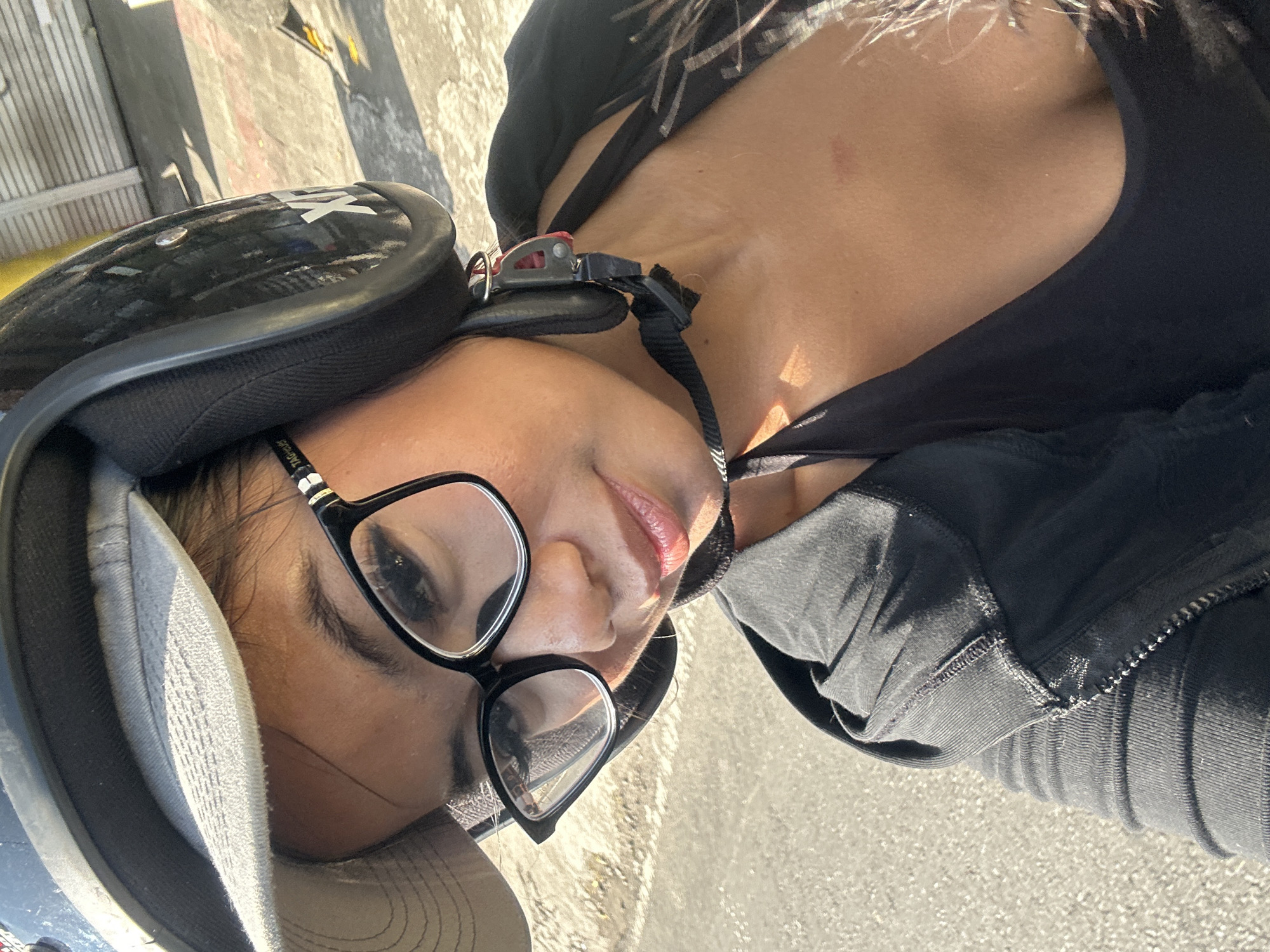 A close-up, selfie-style, eye-level medium shot shows a woman wearing a black motorcycle helmet and a grey cap, as she looks towards the camera. The woman has on black-rimmed glasses and a black tank top under a black hooded jacket. The setting appears to be outdoors on a street in Sukawati, Indonesia, with a sunlit pavement and a white building visible in the background. The lighting suggests it is daytime, with shadows cast on the ground. The woman's skin tone is tan, and she has dark hair.