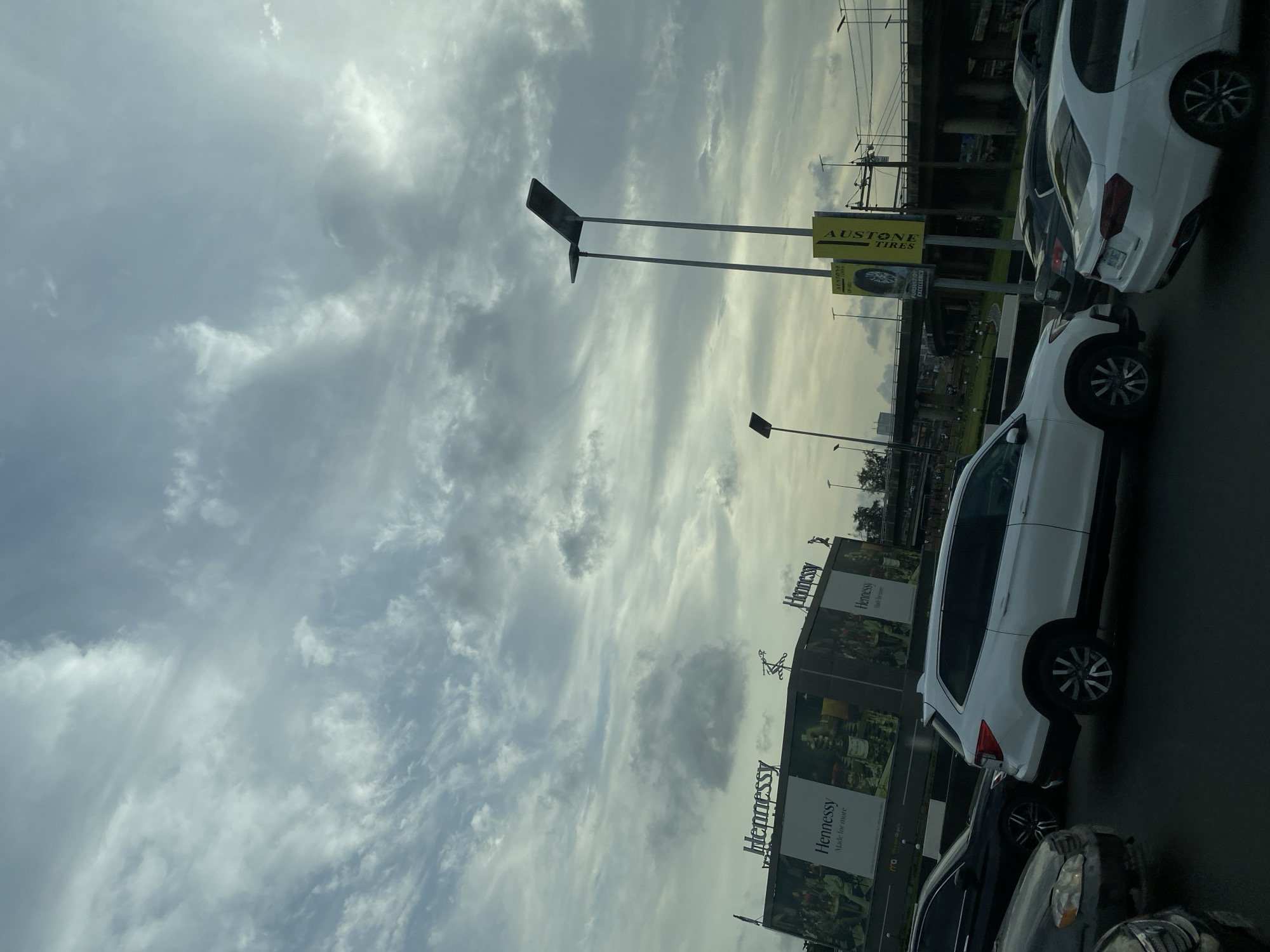 This image, presented with a rotated perspective, depicts a busy urban or highway scene under an overcast sky, consistent with Lagos, Nigeria. The sky is largely covered by diffused grey and white clouds, suggesting either an overcast day or late afternoon/early evening light.
In the foreground and middle ground, several vehicles are visible, indicating traffic or roadside parking. A white SUV or crossover is prominently featured on the right, with a dark car in front of it. Another white sedan is seen further down the road.
The roadside is characterized by significant advertising. A large yellow billboard clearly displays "AUSTONE TIRES" with the slogan "PURSUIT OF EXCELLENCE" and an image of a tire. Nearby, two large billboards prominently feature the brand "Hennessy," accompanied by the tagline "Made for more" and imagery of bottles.
Other urban infrastructure includes multiple tall poles, some appearing to be streetlights or equipped with solar panels. In the background, a concrete overpass or bridge spans across the scene, with overhead power lines visible. Distant structures and sparse greenery can be observed beyond the immediate roadside elements. No individuals are visibly captured in the frame.