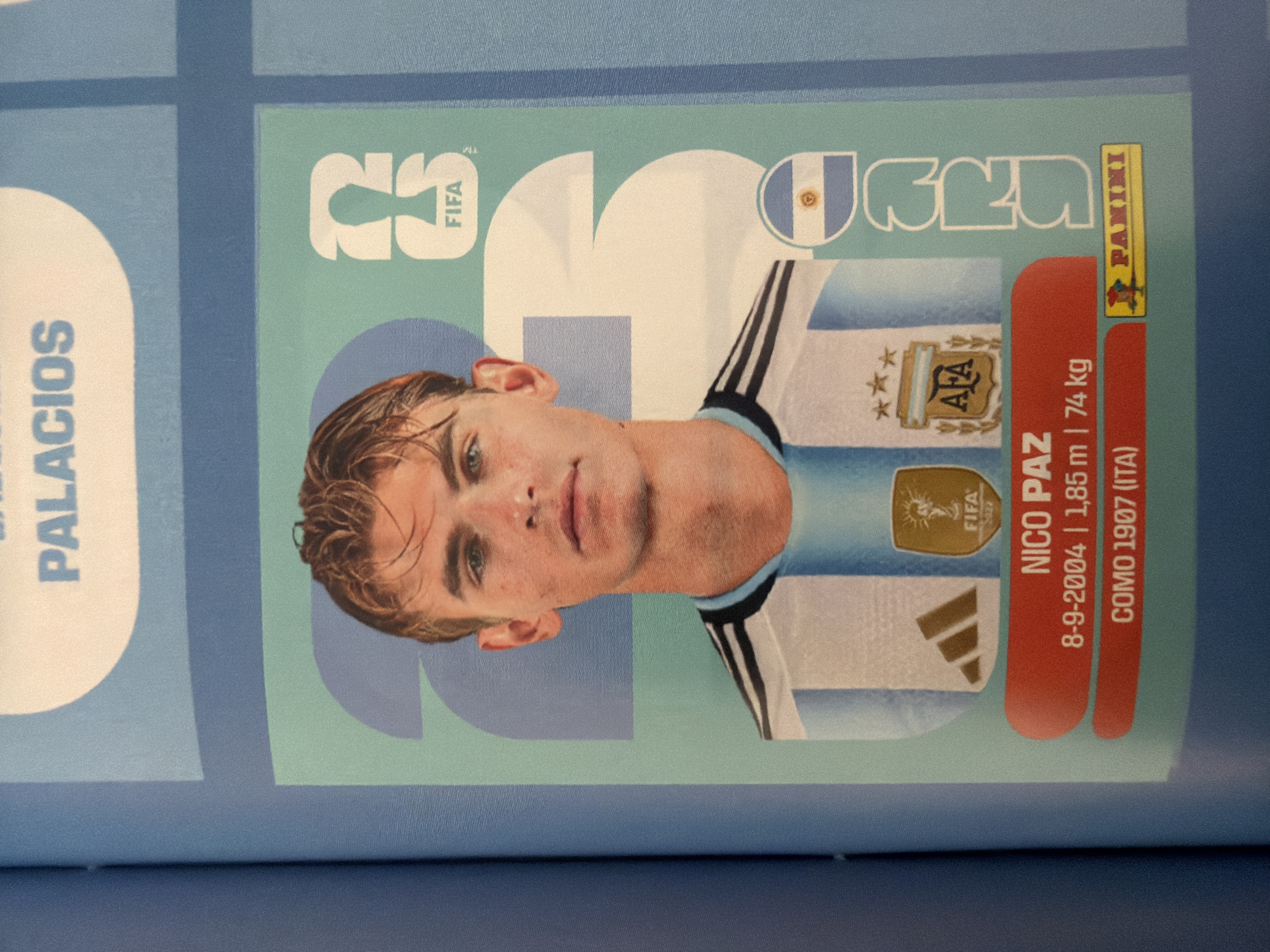 The image is a close-up photograph of a sports trading card. The card features a portrait of a young man with blond hair, wearing an Argentina national football team jersey. The jersey has the team's crest and three stars above it. The player's name, "NICO PAZ," is printed below his image, along with his birthdate, height, and weight: "8-9-2004 | 1.85 m | 74 kg." Below this, it says "COMO 1907 (ITA)." The card also has the FIFA logo and the Panini logo. The background of the card has large, abstract blue and green shapes with the number "23" faintly visible. The overall impression is that this is a collectible item for a football fan.
The image does not contain any information or visual cues that would allow for the determination of its location in the city of Zlatitrap, Bulgaria.