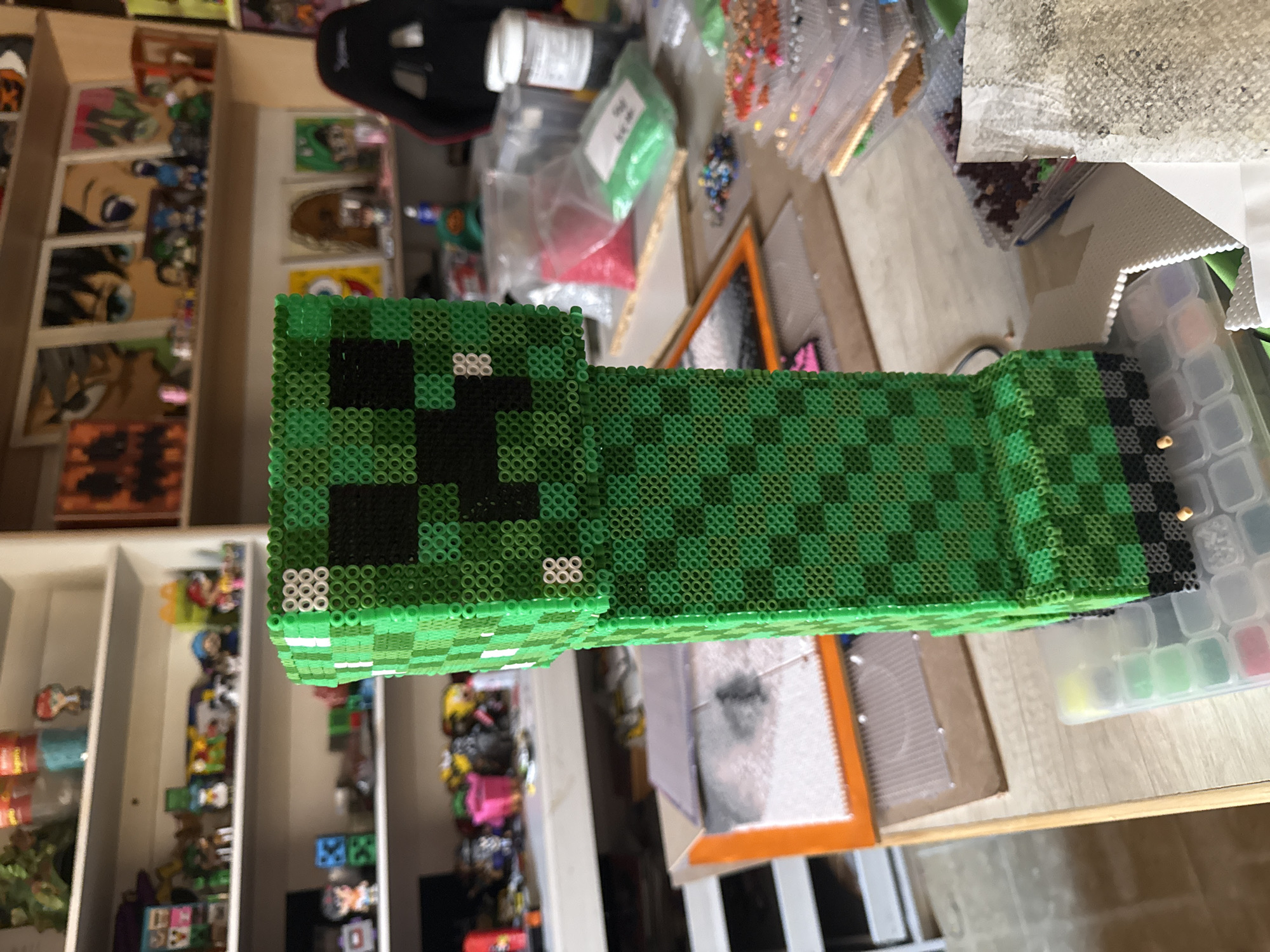 The image shows a 3D Minecraft Creeper made from Perler beads, sitting on a light wooden surface. The Creeper's face is towards the upper-left and its body extends to the lower-right of the image. The Creeper is mostly various shades of green with black details on its face and towards the back end. There are containers and bags with Perler beads, crafting supplies, and an orange-framed picture next to the Creeper. To the left, there is a white shelf unit filled with various objects, including anime portraits and figurines. The setting appears to be a craft or hobby space. The image does not provide the location in the city of Puebla, Mexico.