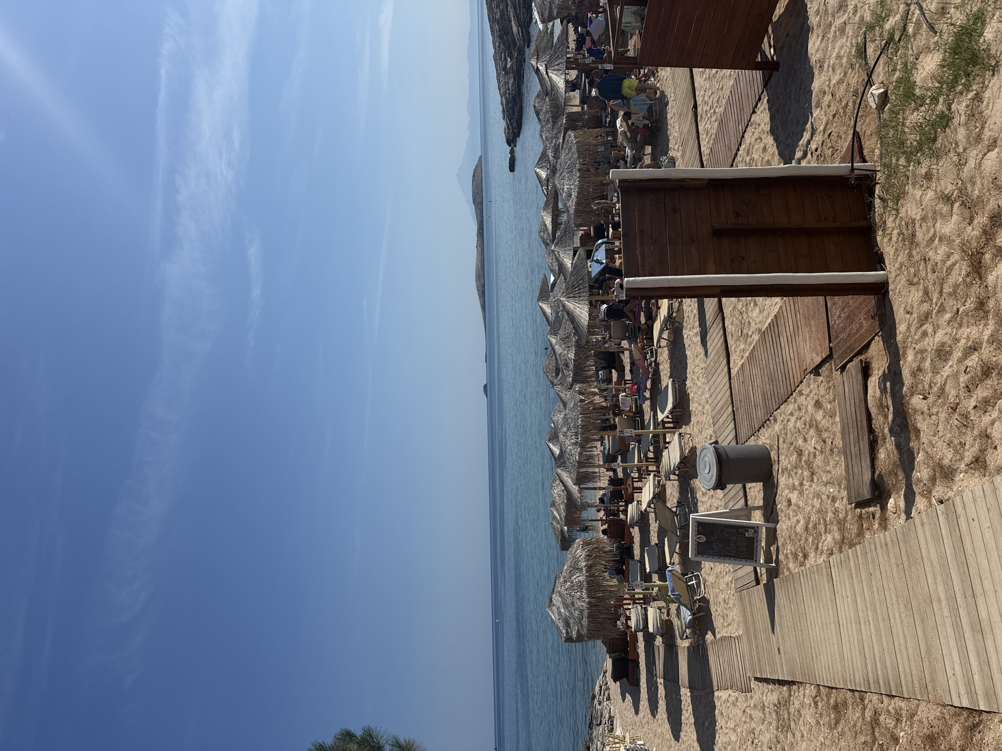 The image depicts a crowded beach in Potos, Greece on a sunny day. The scene is oriented vertically, with the sky occupying the left side of the frame and the beach and sea on the right. Numerous thatched umbrellas and beach chairs are set up on the sand, many occupied by people lounging and enjoying the sun. A wooden boardwalk runs along the beach, providing access to the various seating areas and small wooden structures that appear to be cabanas or service points.
The sea is calm and blue, with a distant coastline visible on the horizon. The sky is clear and bright blue, with a few wispy clouds. The time of day appears to be midday or early afternoon, given the strong sunlight and shadows. The atmosphere seems relaxed and leisurely, characteristic of a popular beach resort.
Visible text is present on a small chalkboard near a trash can and a wooden bench. The text on the chalkboard is difficult to read clearly due to the image resolution, but it appears to be a menu or a sign with handwritten content in a light color against a dark background. There are also some numbers visible on the tables set up under the umbrellas, likely indicating table numbers. The overall impression is a vibrant and well-utilized beach setting, typical of a summer vacation spot in Greece.