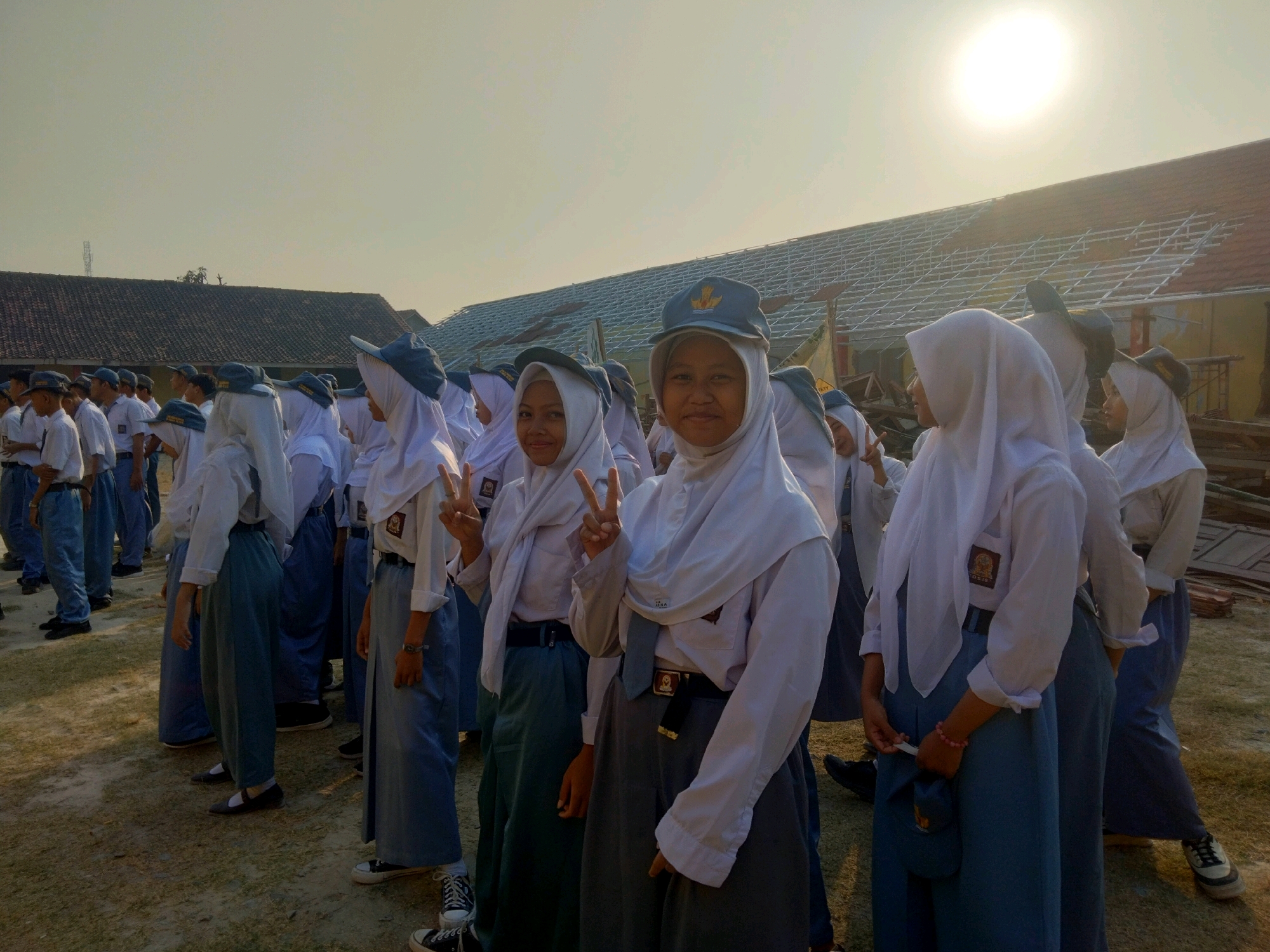 In Suka Sari, Indonesia, a group of students in school uniforms are gathered outdoors. The students are wearing white shirts, blue skirts or pants, and grey caps. Some of the female students are also wearing white hijabs. They appear to be standing in formation, possibly for a school event or assembly. The background shows buildings with tiled roofs, some of which appear to be under construction or renovation. The sun is visible in the sky, casting a bright light and creating a warm, hazy atmosphere. The ground is covered in dirt and gravel.
