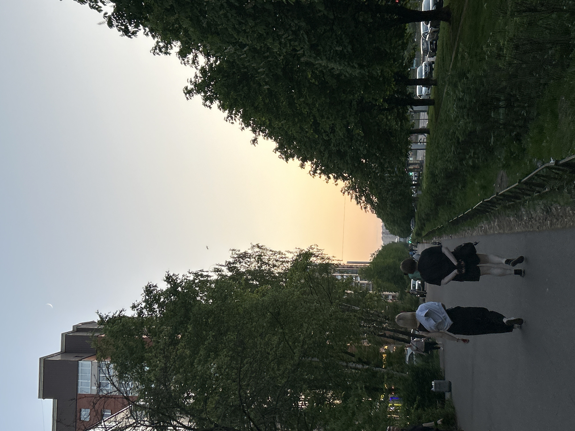 The image depicts a pathway in Saint Petersburg, Russia, lined with trees. Two individuals are walking on the path; one is wearing a white top, the other a black top. A building is visible on the left side of the frame. In the background, cars are parked along a street. The sky is bright with a sunset or sunrise. A crescent moon is visible in the sky.