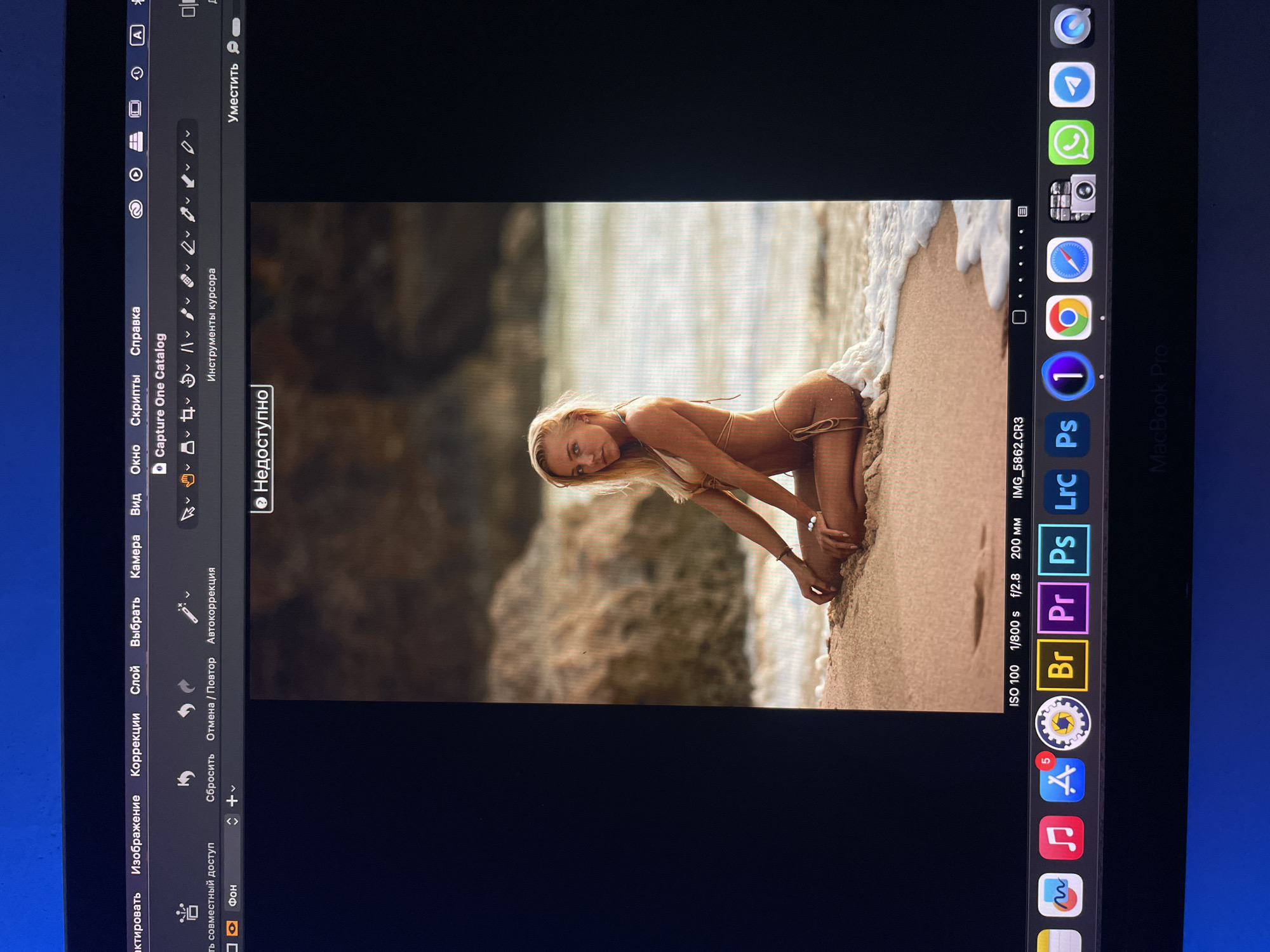 The image is a photograph of a woman kneeling on a sandy beach near the water's edge. She is wearing a tan bikini and leaning forward with her hands on the sand. There is a rocky cliff or land formation in the background on the left.
The image is displayed on the screen of a Macbook Pro. Visible in the screenshot are the Capture One editing software interface and icons on the Mac's dock, including Safari, Chrome, Finder, Capture One, Photoshop, Premiere Pro, Bridge, System Settings, App Store, Music, Notes, and others. Also visible in the dock are icons for Telegram and WhatsApp. The operating system language is Russian. The ISO, shutter speed, aperture, and focal length of the original photograph are also visible.
The image does not include any location information.