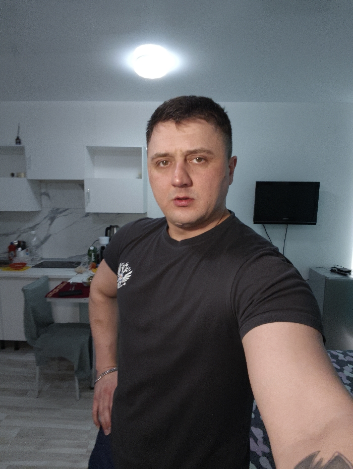 This image displays an adult male subject positioned indoors within a residential setting, presented as being in the city of Совхозный, Russia. The male, appearing to be in his 30s or 40s, has short, dark hair and a neutral facial expression while looking directly at the camera. He is wearing a dark-colored, short-sleeved t-shirt featuring a white and black emblem, resembling a double-headed eagle, on the left chest. A silver-colored chain bracelet is visible on his left wrist, and a dark-colored tattoo is partially visible on his right forearm. His left hand is placed on his hip, and his right arm is extended, holding the device used to capture the image.
The background reveals a combined kitchen and living area. On the left, a modern kitchen setup includes white upper and lower cabinets, a light-colored countertop with a veined pattern, and a white range hood. A dark-colored table with two chairs covered in light grey fabric slipcovers is situated in front of the kitchen area. The countertop holds various objects, including bottles and a kettle. On the right, a small black flat-screen television is mounted on a light-colored wall above a light-colored metallic cabinet. A patterned fabric, possibly part of a bed or seating, is visible in the lower right quadrant. The room features light-colored walls and ceiling, with a single illuminated circular ceiling light fixture. The flooring consists of light-colored wooden or laminate planks.