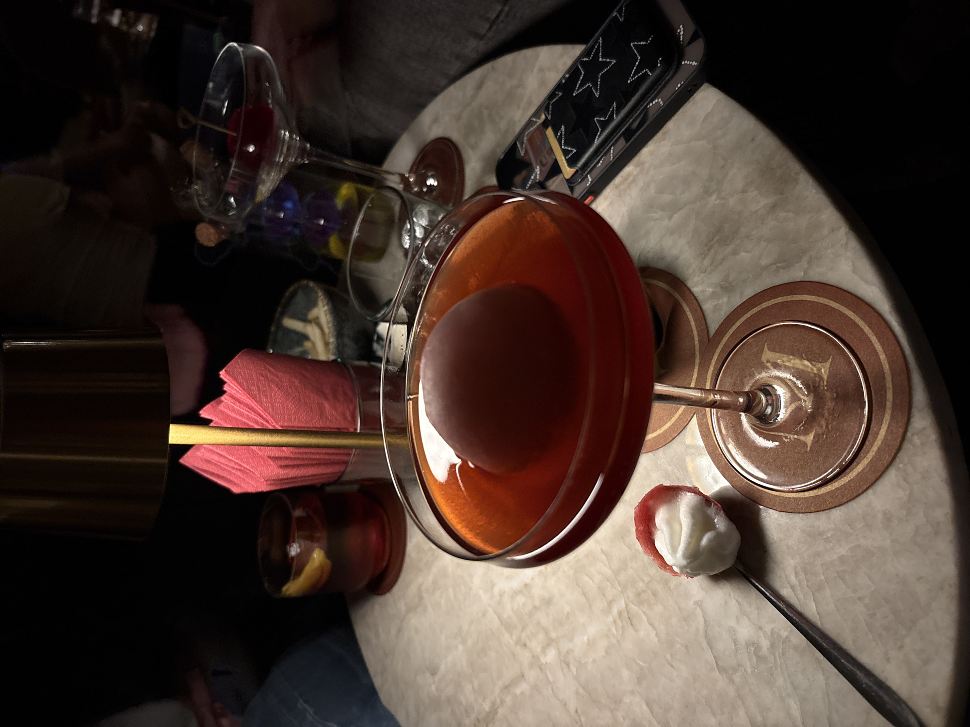 The image depicts a cocktail bar setting in what is speculated to be Mexico City, Mexico. Centered is a cocktail glass filled with a reddish-brown liquid and containing a large spherical ice ball. The glass rests on a circular coaster with a brown rim and an indistinct emblem. To the right of the glass is a spoonful of white substance, appearing to be either whipped cream or a meringue, on a spoon. Arranged around the cocktail glass are other bar accessories, including a gold-colored metal shaker, folded red napkins, an additional cocktail glass, and a container with a star pattern. The surface appears to be marble or a similar polished stone. The background is dimly lit, emphasizing the focus on the drinks and accessories.