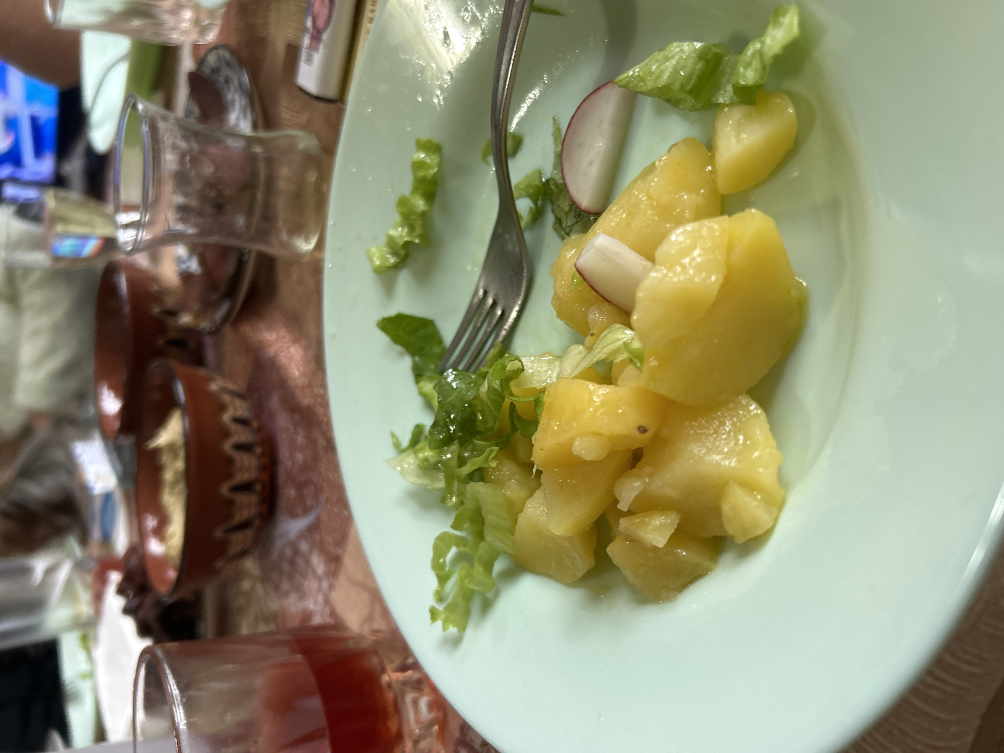 A tilted, close-up shot shows a light green plate containing a potato salad with lettuce and sliced radish. A silver fork rests on the plate among the salad. In the blurred background, several drinking glasses are visible, along with a decorative bowl. The scene appears to be a table setting indoors, possibly a home, in Polski Izvor, Bulgaria. The setting seems informal. There are no people clearly visible in the image.