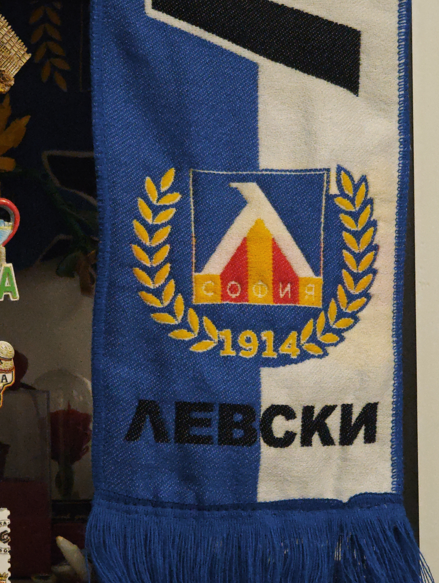 A close-up shot depicts a Levski Sofia football scarf. The scarf is divided vertically in blue and white with a possible small area of black at the top. On the blue section, there's a logo featuring a triangle in red, yellow, and white, surrounded by a laurel wreath. The text "СОФИЯ" (Sofia) and "1914" are visible within the wreath. Below the logo, the word "ЛЕВСКИ" (Levski) is written in large black letters, crossing the blue and white sections. The scarf has blue tassels at the bottom. The context suggests a display or collection, with other items visible out of focus in the background. The text and logo confirm the location is related to Levski Sofia, a football club in Sofia, Bulgaria.