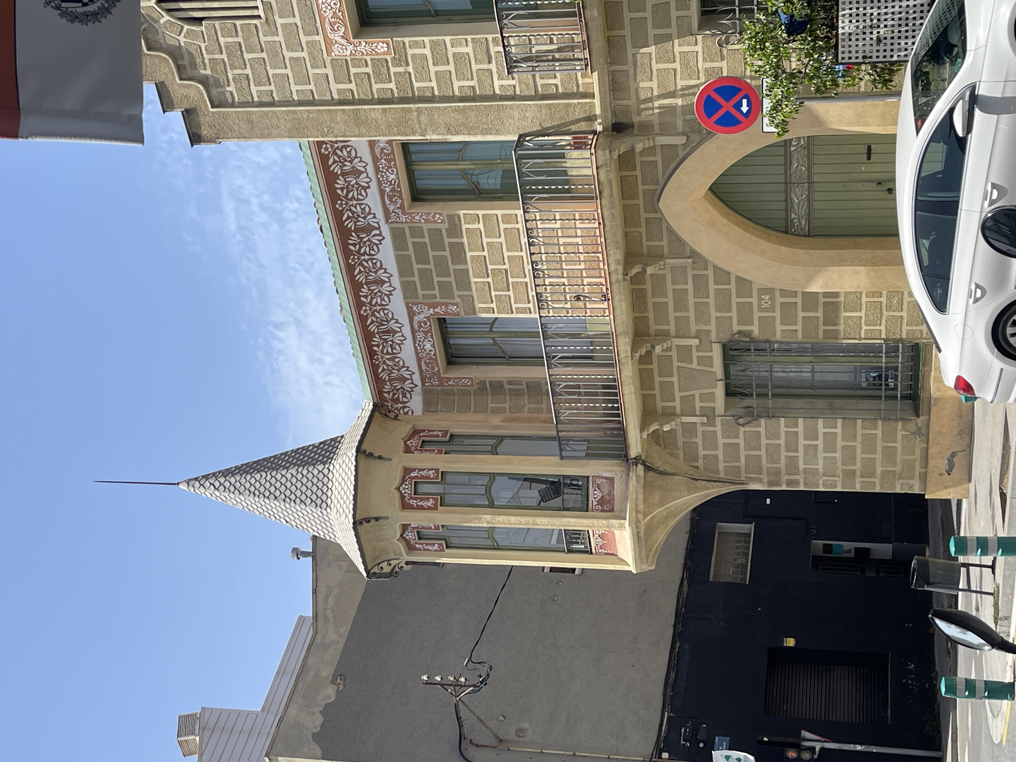 The image shows a street view of a building with distinct architectural features in Badalona, Spain. The building is characterized by its light-colored brick facade, decorative floral patterns along the upper sections, and a prominent pointed tower with a tiled roof. There are several windows, some of which are bay windows, and balconies with ornate metal railings. A large arched doorway with green double doors is visible at the base. The street in front of the building features a white car, traffic lights, bollards, and a "no parking" sign with an arrow. Adjacent to the main building is a darker, more modern-looking structure with horizontal blinds on a window. The sky is blue with scattered clouds, indicating a clear day. The overall scene suggests an urban environment with a blend of older, decorative architecture and more contemporary elements.