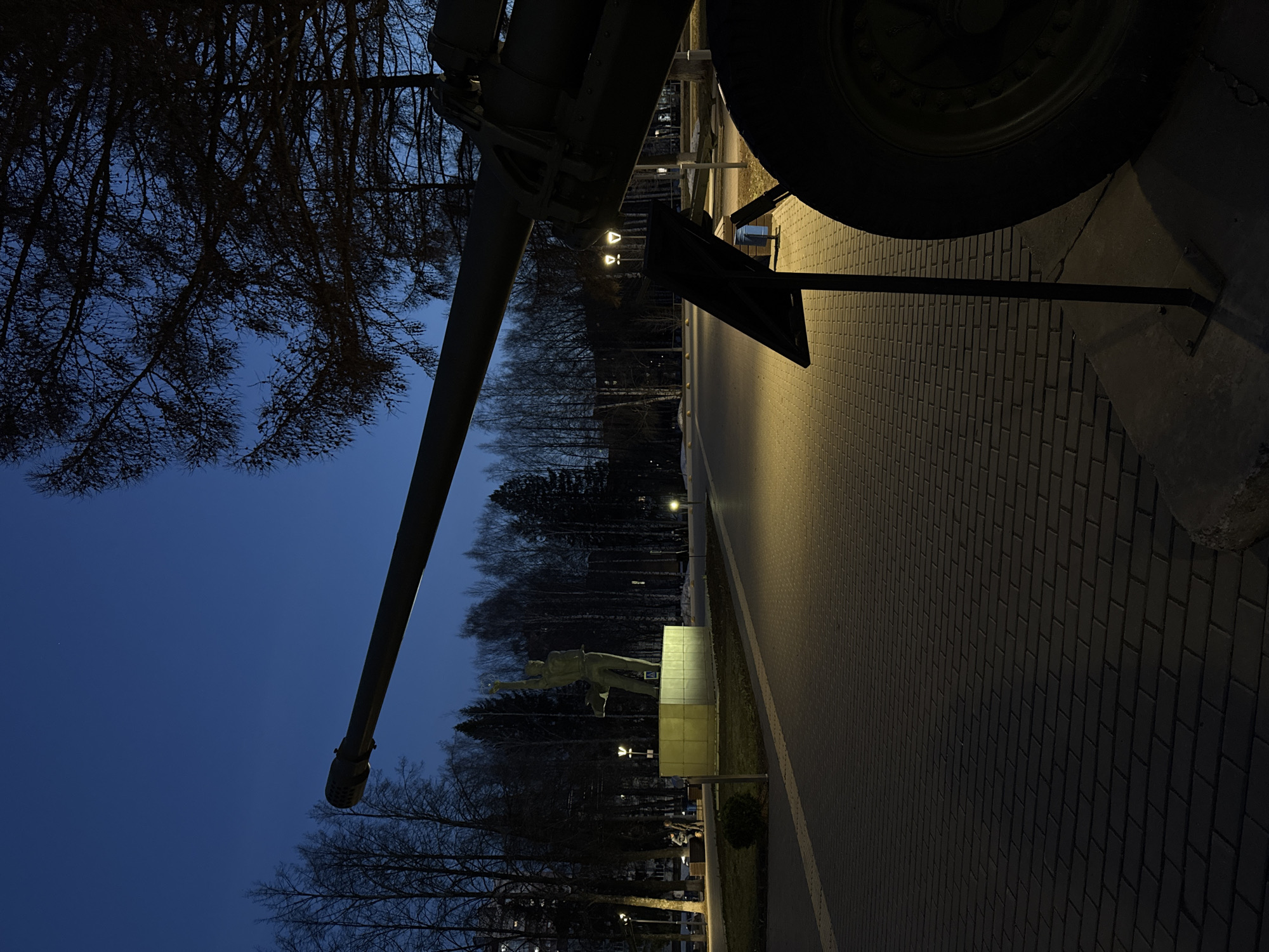 This photo, taken at night, shows a cannon pointed skyward near a statue of a man, likely a monument in a park in Izhevsk, Russia. The cannon is dark green and positioned to the left of the frame, with the end of the barrel extending into the blue sky. The tire of the cannon and a black brace cast shadows on the gray brick pathway. In the background, bare trees are visible. Beyond the statue, the pathway stretches into the distance, illuminated by streetlights.