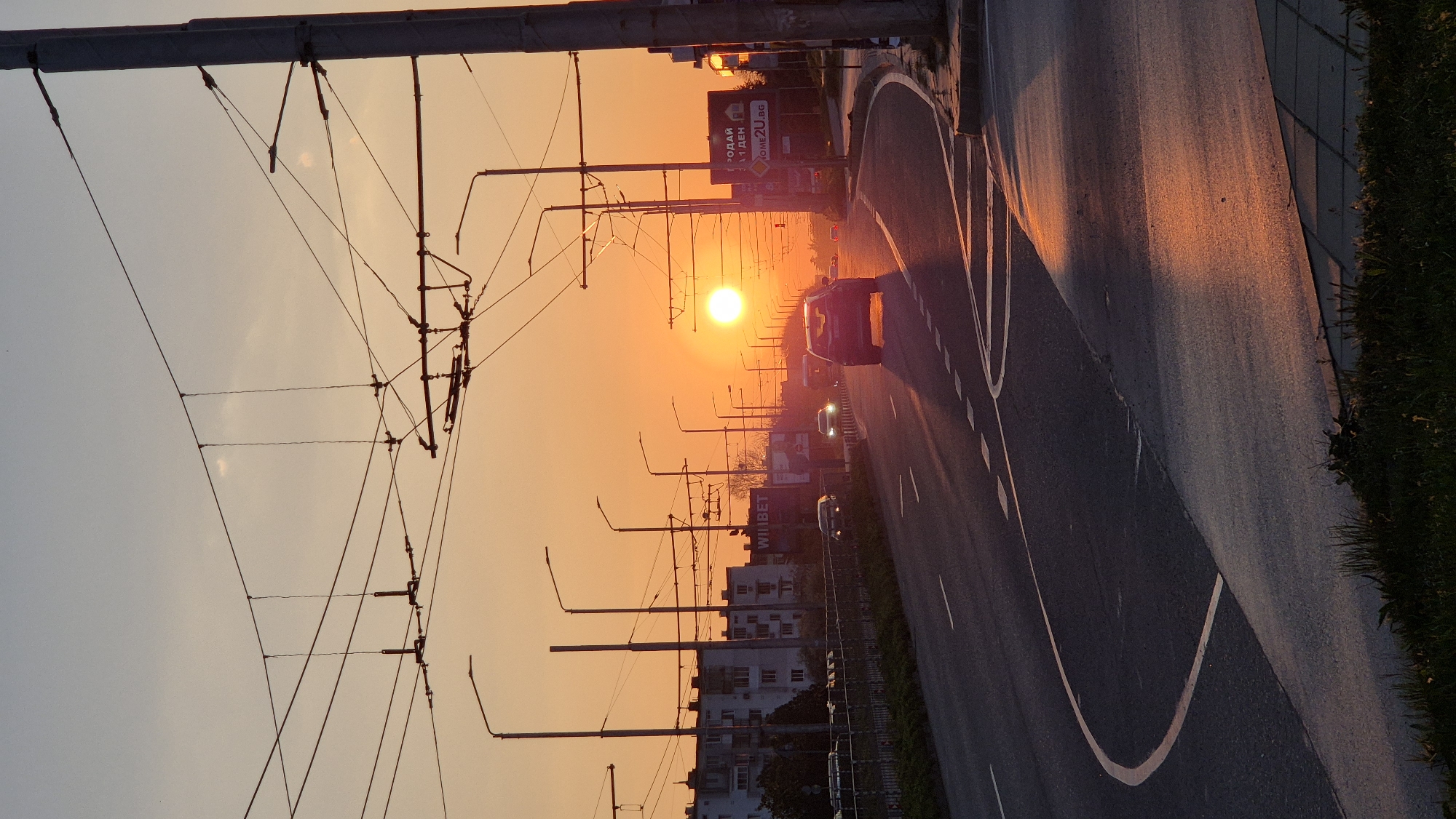 The image shows a street in Varna, Bulgaria, at sunset. The sun is positioned directly in the center of the frame, creating a strong focal point and casting a warm orange glow. Utility poles and wires line the left side of the street, creating a linear pattern. A road with white markings is on the right, with cars driving on it. Buildings are visible in the background. The sky is a gradient of colors, ranging from a lighter orange near the horizon to a pale blue higher up.