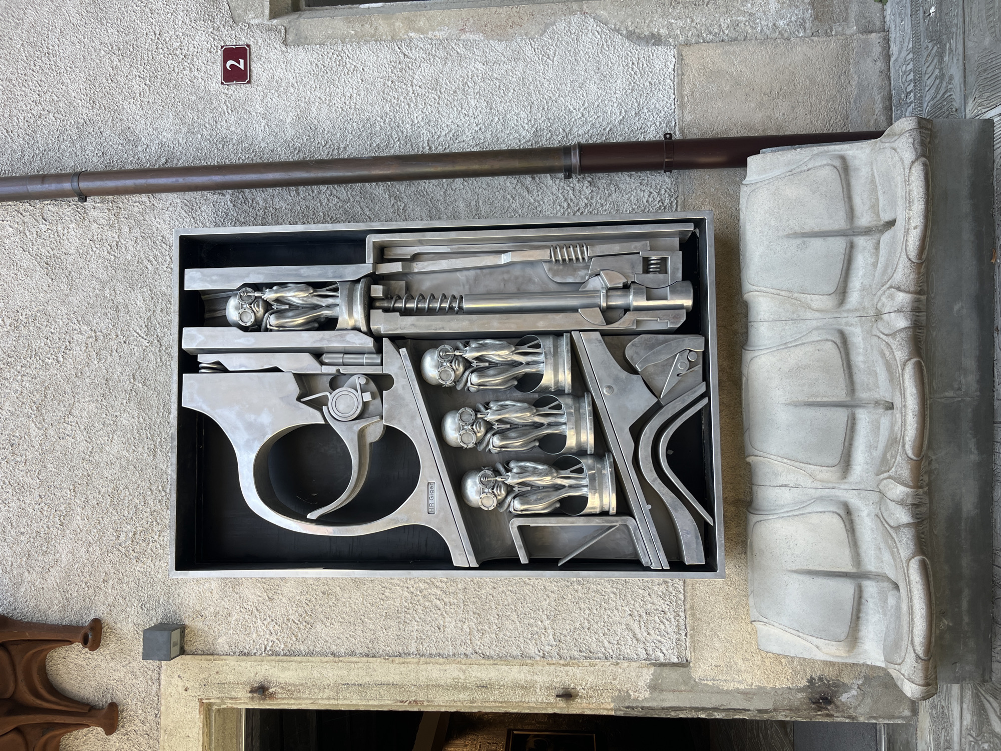 This is a medium shot of a public art installation in Epagny, Switzerland. The installation consists of a large, metallic sculpture resembling a gun mechanism, with three small, metallic figures wearing goggles and appearing to be in a fetal position, positioned within the sculpture. Below the sculpture is a grey, molded bench with a textured surface, designed to seat multiple people. The sculpture is mounted on a textured grey wall, to the right of which is a dark brown downspout. A rectangular, maroon sign with the number "2" in white is affixed to the wall above the downspout. To the left of the sculpture, a building entrance with a visible doorway and frame is present, with a small, dark grey light fixture mounted on the wall above the entrance. The lighting suggests it is daytime.
