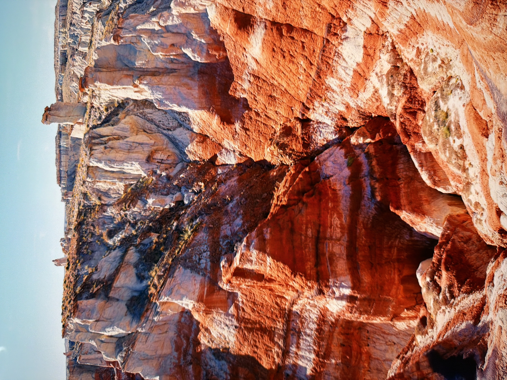 This is a high-angle, full shot of a rugged, arid canyon landscape under a clear, bright blue sky. The canyon walls are characterized by layered rock formations in vibrant shades of orange, red, and white, exhibiting a dramatic striated pattern. The terrain is steep and eroded, with sharp vertical spires and formations dotting the upper ridges. Sparse, dry vegetation, appearing as tufts of grass, grows on some of the flatter surfaces. The lighting suggests a sunny day, casting shadows that accentuate the textures and depth of the rock formations. The scene depicts a natural geological formation. There are no people, man-made objects, or signs of urban life. This landscape is not located in Kyiv, Ukraine; it is a natural geological formation.