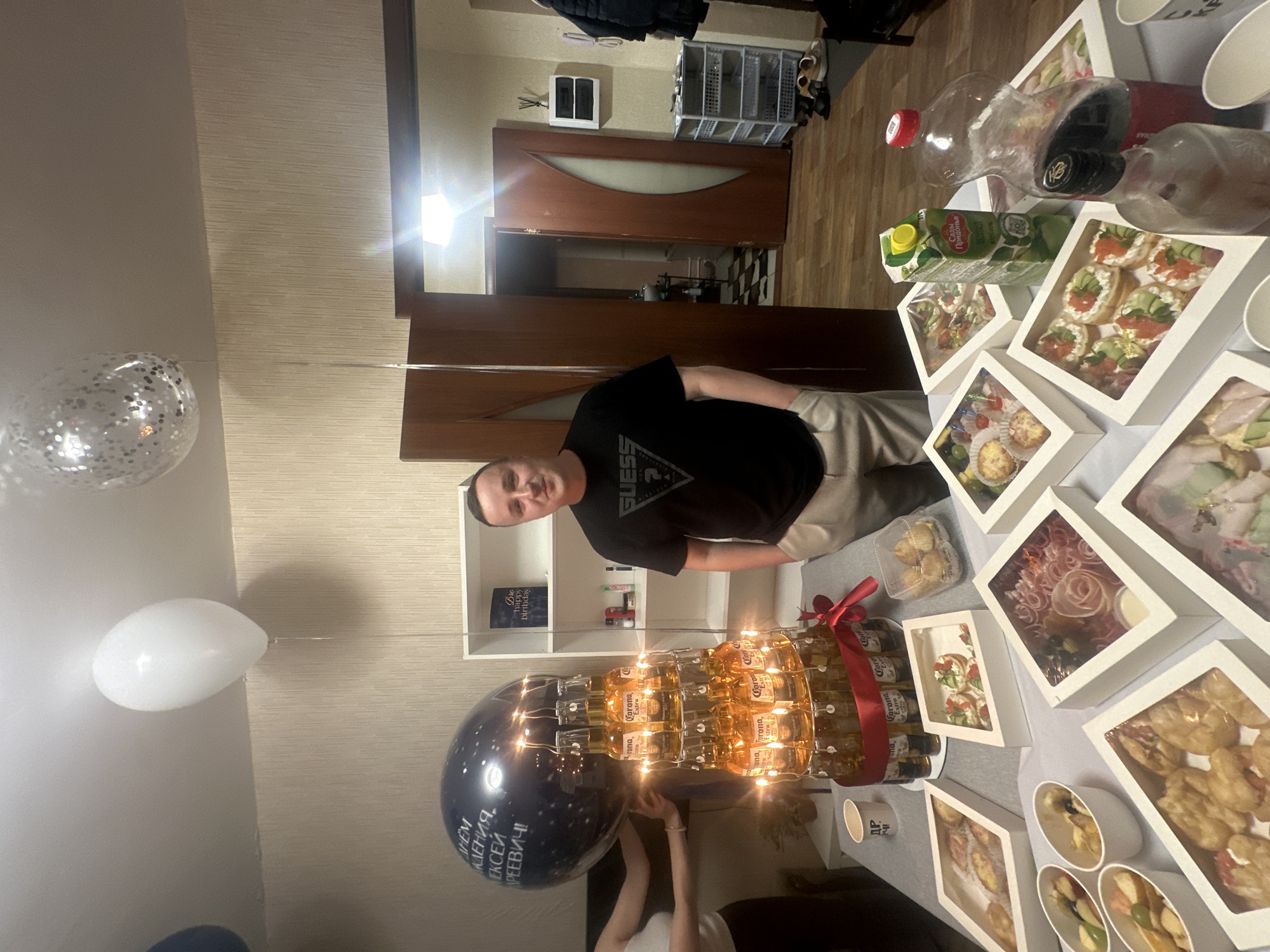 The image depicts an indoor scene, likely a domestic setting, with a table laden with food and drinks. A man, wearing a black t-shirt with a "Guess" logo and light-colored pants, stands near the center of the frame, facing the camera with a slight smile. To his left, a tower of Corona beer bottles, illuminated by string lights, is decorated with a red ribbon. A large, dark blue balloon with white lettering is partially visible behind the beer tower. The table also displays various food containers, some filled with appetizers and snacks, a carton of juice, and bottles of drinks. In the background, a doorway leads to another room, and shelves are visible with decorative items. The overall impression is of a celebration or gathering. No specific landmarks or contextual clues in the image indicate the city of Perm, Russia.