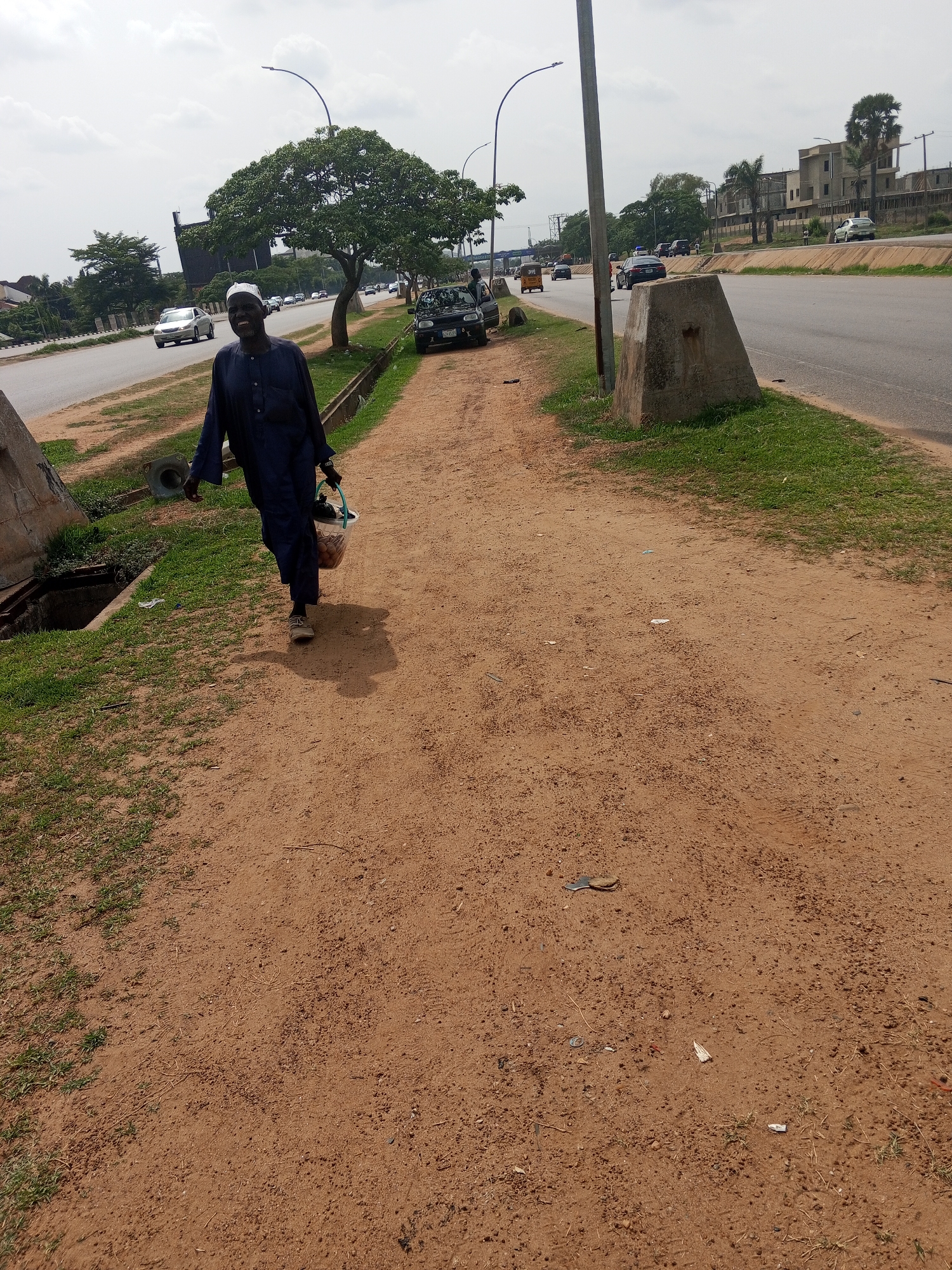 A man dressed in dark blue clothing and a white cap walks along a dirt path. He is carrying a bucket in his left hand. The path runs parallel to a major road with several cars driving on it. The scene is in Abuja, Nigeria, based on the road markings and building styles. There are concrete barriers and light posts along the road. Grass grows between the path and the road. A tree and some other greenery are visible in the background.