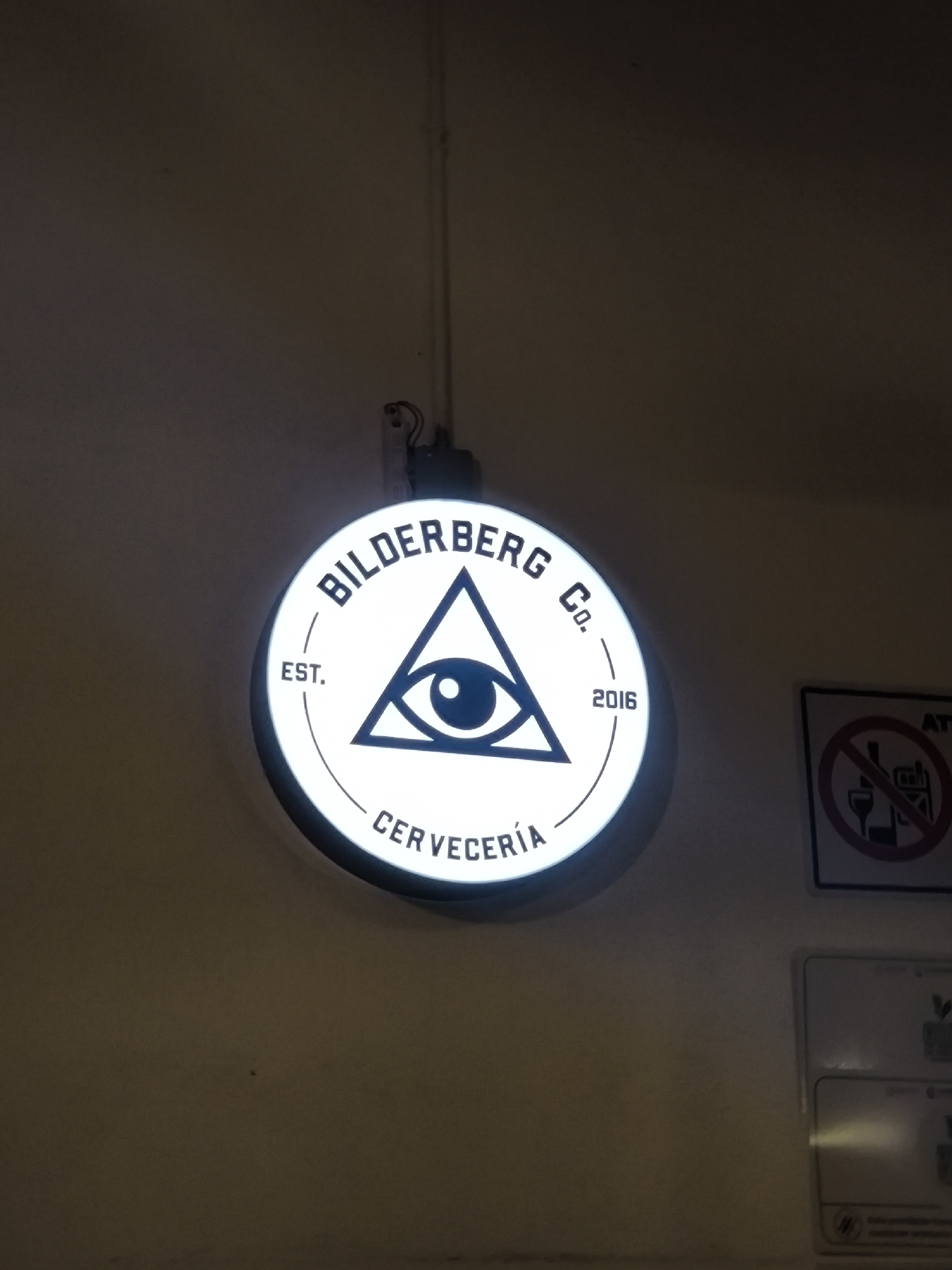 Here's a concise description of the image:
The image shows a circular illuminated sign hanging on a wall. The sign reads "BILDERBERG Co." at the top and "CERVECERÍA" (brewery) at the bottom, with "EST. 2016" on either side of a central emblem: a stylized eye within a triangle. To the right of the sign, partially visible, are two rectangular signs; the upper sign contains a graphic showing a circle with a line through it superimposed over images of drinking glasses, implying a ban on alcohol, and the sign below appears to be informational or regulatory. The wall is painted in a plain, neutral color, and a hanging wire and mounting box are visible above the sign. Without additional contextual information from the image, the exact location in Puebla, Mexico cannot be determined.