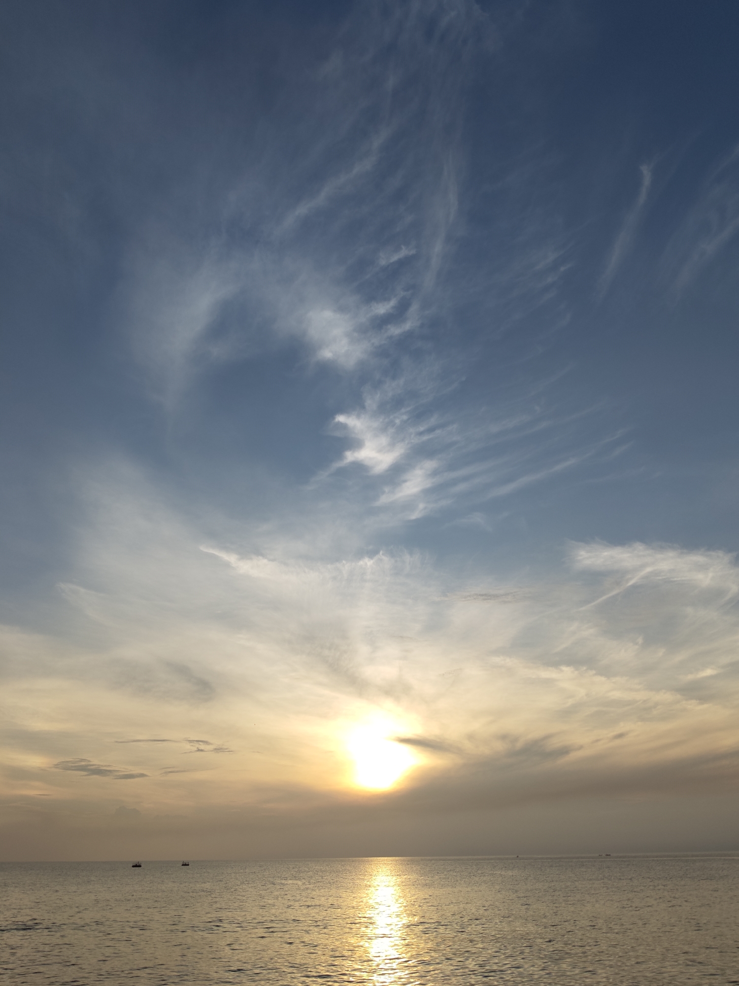 This image captures a serene sunset over the waters of Lhokseumawe, Indonesia. The sky is a gradient of blue, transitioning to soft yellows and oranges near the horizon where the sun is setting. Wispy white clouds streak across the sky, adding texture and depth to the scene. The sun itself is a bright, diffused orb, casting a shimmering golden reflection on the calm sea below. A few small boats are visible in the distance, dotting the vast expanse of water. The overall impression is one of tranquility and natural beauty.
