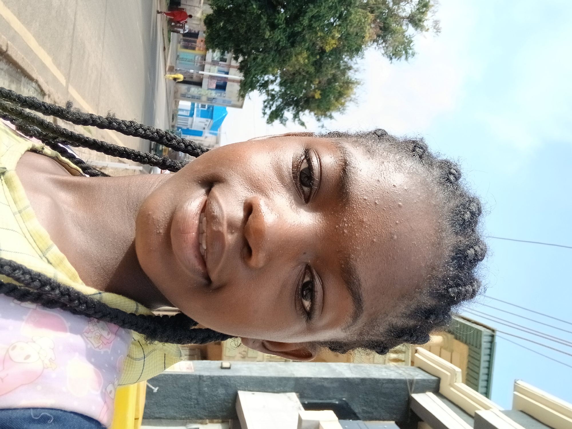 A medium close-up photograph, originally rotated, shows a young Black woman smiling warmly at the camera. Her face, positioned slightly to the right of the frame, features clear, dark eyes and a broad, open-mouthed smile, revealing teeth. Her dark skin appears somewhat shiny, possibly from sunlight or perspiration. She has dark braided hair, which frames her face. She is wearing a light-colored, possibly yellow, top with a textured pattern, and another garment with a pastel-colored, possibly cartoon-patterned design, is visible underneath.
The background reveals an outdoor, urban or semi-urban setting under bright daylight, suggesting mid-day. Behind the woman, a paved road with a yellow line is visible. In the distance, there are various buildings, some with blue, white, and beige facades. A large tree with dense green foliage stands towards the background. Faint figures, including a person in a red shirt and another in yellow, are visible further down the road. Overhead, power lines or cables crisscross against a clear, light blue sky with some soft white clouds. The overall impression is a casual, sunny day in Jos, Nigeria. There is no visible text in the image.