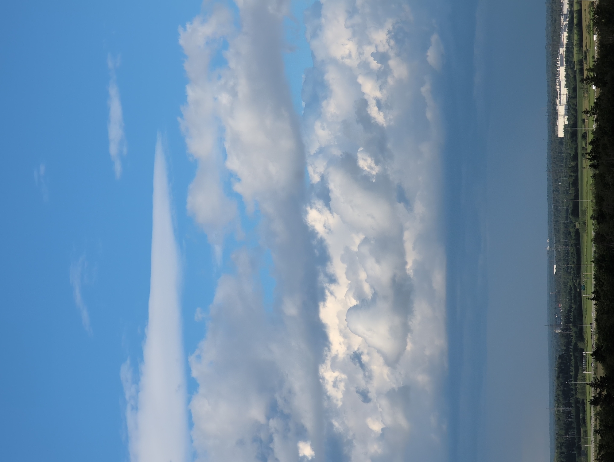 The image depicts a sky with varying cloud formations and a distant landscape. The sky is predominantly blue, with scattered white and gray clouds. Some clouds are wispy and elongated, while others are more dense and textured. Towards the right side of the image, the sky appears darker, suggesting a potential storm or overcast conditions. Below the sky, a landscape is visible, characterized by a line of trees and buildings. The buildings appear to be part of an industrial or commercial area, with several white structures visible. A road or highway runs along the edge of the landscape, with streetlights lining it. The presence of trees suggests a semi-urban or suburban environment. The overall scene is calm, with no discernible activity or people. The lighting suggests it is daytime, possibly late afternoon or early evening, given the contrast in cloud darkness. The location context provided places this scene in Saint Petersburg, Russia, though no specific landmarks are identifiable. There is no visible text in the image.