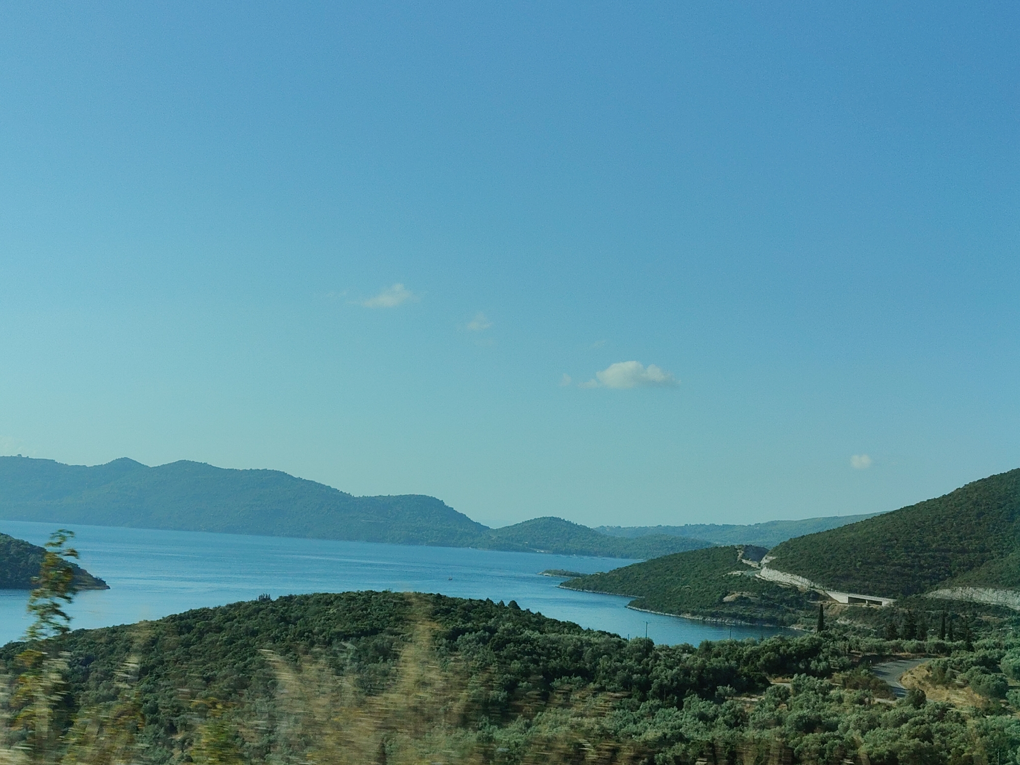 This image depicts a scenic view of the Municipal Unit of Pelasgia, Greece, likely taken from an elevated vantage point. The foreground is dominated by lush, green vegetation, characterized by dense trees and shrubs, with some appearing slightly blurred, suggesting movement such as a vehicle passing by. This foliage covers rolling hills that slope down towards a body of water.
In the middle ground, a calm, blue sea or large lake is visible, with several verdant islands or peninsulas jutting out into it. The coastline is irregular, creating coves and inlets. In the distance, a range of larger, mountainous islands or landmasses form a hazy backdrop against the horizon. The sky is a clear, pale blue with a few wispy white clouds scattered across it.
A paved road with guardrails is visible winding its way up the side of one of the hills in the right mid-ground. There are also some structures, possibly buildings or infrastructure related to the road, visible on the hillside. The overall impression is one of a serene, natural landscape with a touch of human development in the form of the road. There are no people visible in the image.