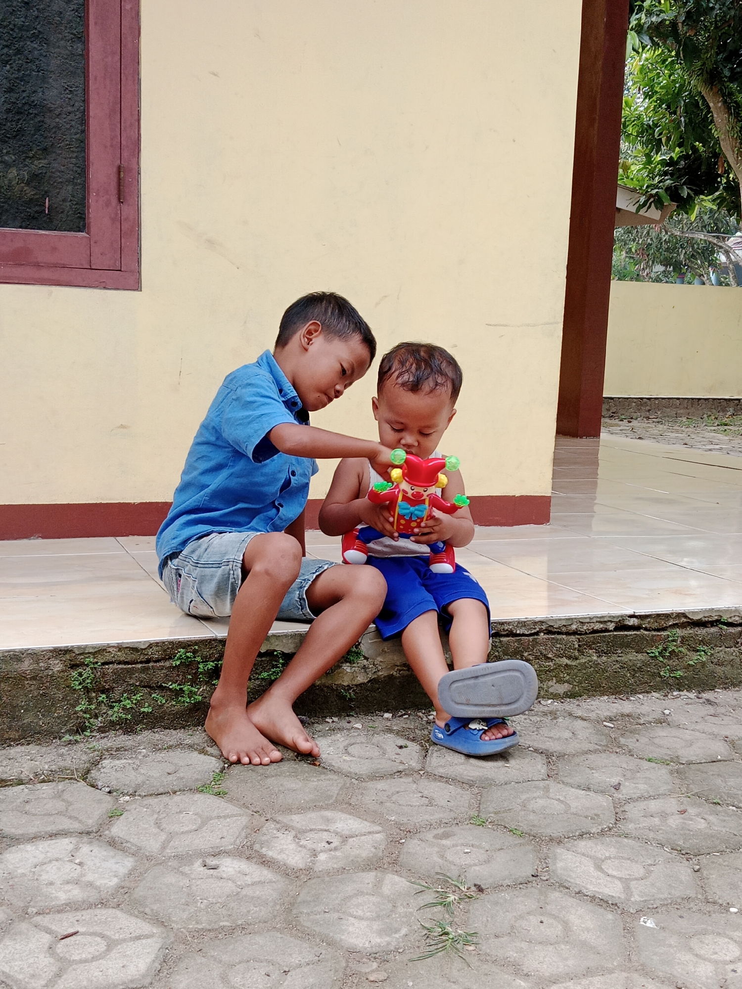 Two young boys are sitting on a tiled ledge outside a building in Parage, Indonesia. The older boy, on the left, wears a blue short-sleeved shirt and denim shorts. He is looking down at the toy in his younger brother's hands. The younger boy, on the right, wears a white tank top and blue shorts. He holds a colorful toy clown with green balls on its head and red shoes. The older boy's bare feet are visible, while the younger boy wears blue sandals. The building behind them is painted a pale yellow with a dark brown trim and a dark red-framed window on the left. The ground in front of the ledge is paved with hexagonal concrete blocks. Green plants are growing along the edge of the ledge and between some of the paving stones. Trees and foliage are visible in the background to the right. The scene appears to be a casual outdoor setting.
