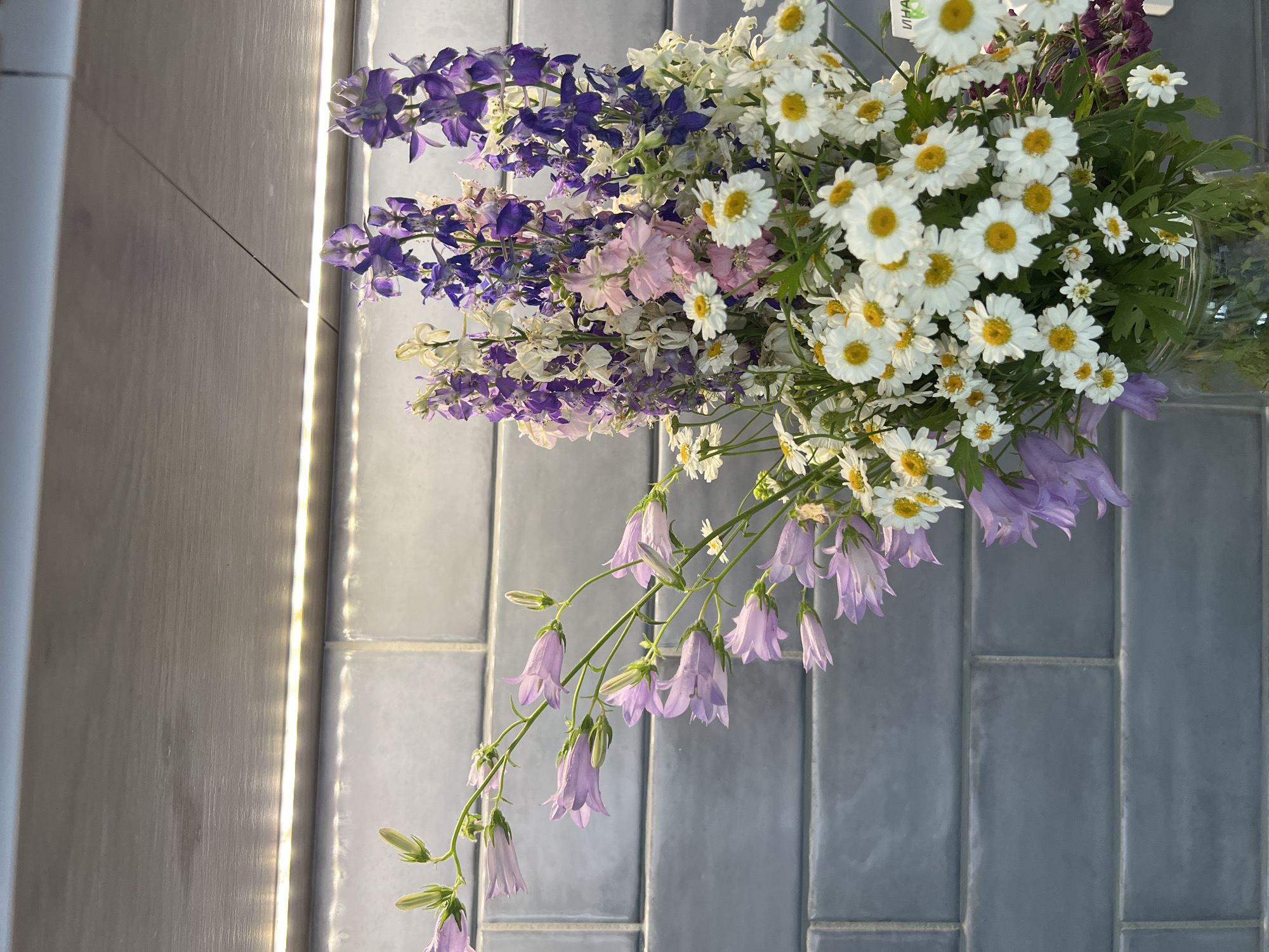 A close-up, vertical shot shows a bouquet of flowers in a glass vase against a grey tiled wall. The bouquet contains several types of flowers: lavender-colored bell-shaped flowers hang on delicate stems, interspersed with purple and pink delphinium spikes, and numerous small white daisies with yellow centers. The wall behind the flowers is composed of rectangular grey tiles, arranged horizontally. A strip of bright white LED lighting is visible along the top edge of the tiled wall, casting a subtle glow. The overall impression is one of natural beauty captured in a well-lit indoor setting.
