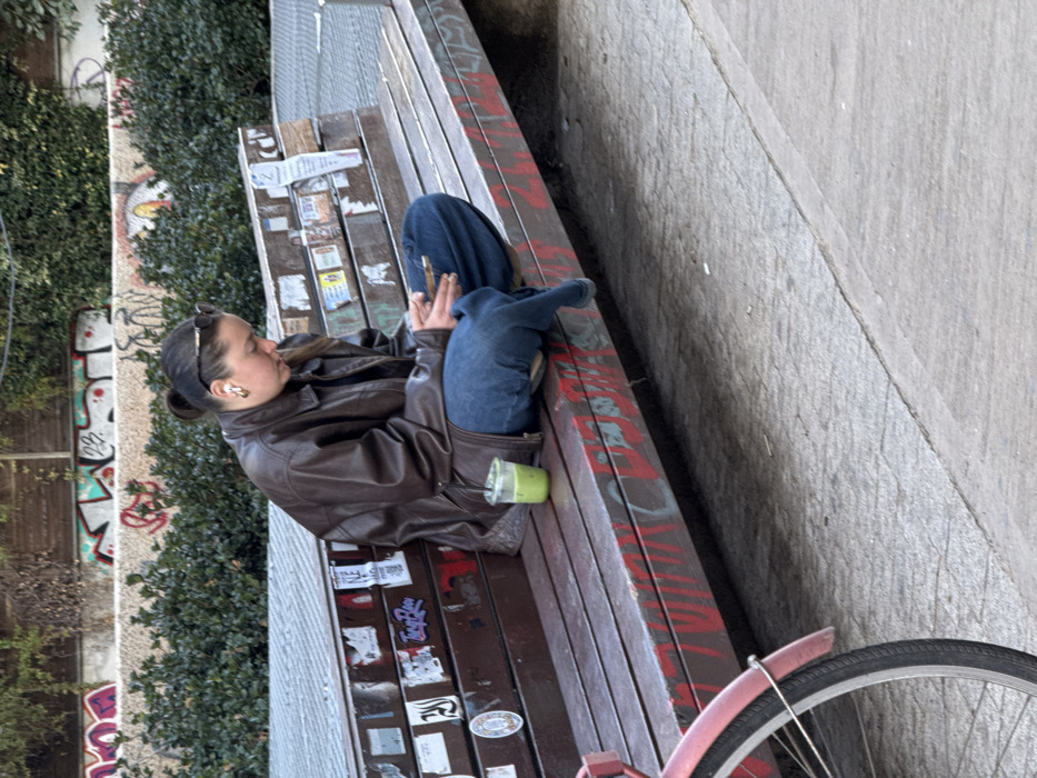 A young adult female is seated on a multi-plank wooden bench in an urban public space in Tel-Aviv, Israel. She is positioned with her legs crossed at the ankles, facing towards the right side of the frame, with her head angled downwards. She wears a dark brown leather jacket, a dark top, and loose blue jeans. Sunglasses rest on top of her head, and white wireless earbuds are visible in her ears, along with large gold-colored hoop earrings. Her hands hold a light-colored smartphone, at which her gaze is directed.
To her left, on the bench surface, a clear plastic cup containing a light green liquid and a dark straw is present. The wooden bench planks are covered with numerous stickers and red spray paint graffiti along the lower front edge. The immediate ground in front of the bench consists of light grey paving stones. In the lower-left foreground, the front wheel and fender of a red bicycle are partially visible. The background features a dense row of green bushes, behind which a concrete wall is heavily covered in multi-colored graffiti tags and writing.
