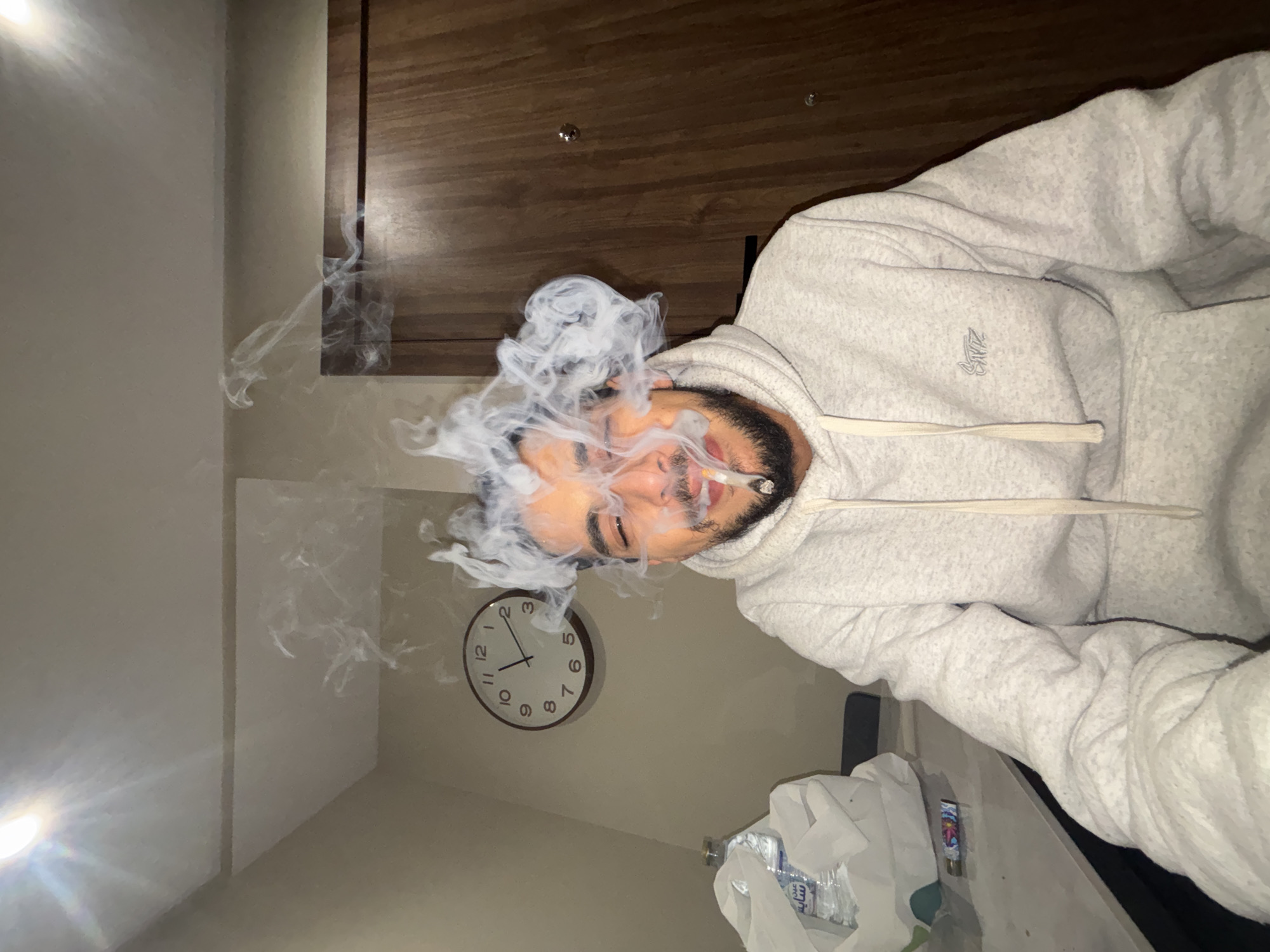 This image depicts a male subject indoors, located in the city of Tanger, Morocco. The subject, visible from the chest up, has dark hair, a beard, and a mustache. He is wearing a light grey hooded sweatshirt with white drawstrings and a dark grey embroidered logo on the left chest area. He is holding a lit hand-rolled cigarette in his mouth, from which a large volume of white smoke billows upwards and around his head, partially obscuring his face and causing his eyes to appear squinted. He exhibits a slight smile.
The immediate environment includes a light-colored wall behind the subject. To his left, an analog wall clock with a dark rim and white face shows the time as approximately 11:25. Below the clock, on a light-colored table or counter surface, a white plastic bag contains a clear plastic water bottle with a blue label featuring Arabic text. A colorful, patterned lighter rests on the same surface. To the subject's right, a wooden door with vertical grain is visible, featuring two circular metallic door handles/knobs. A bright, recessed light fixture is active on the ceiling in the upper left portion of the frame, casting a glare. The overall lighting appears artificial and warm.