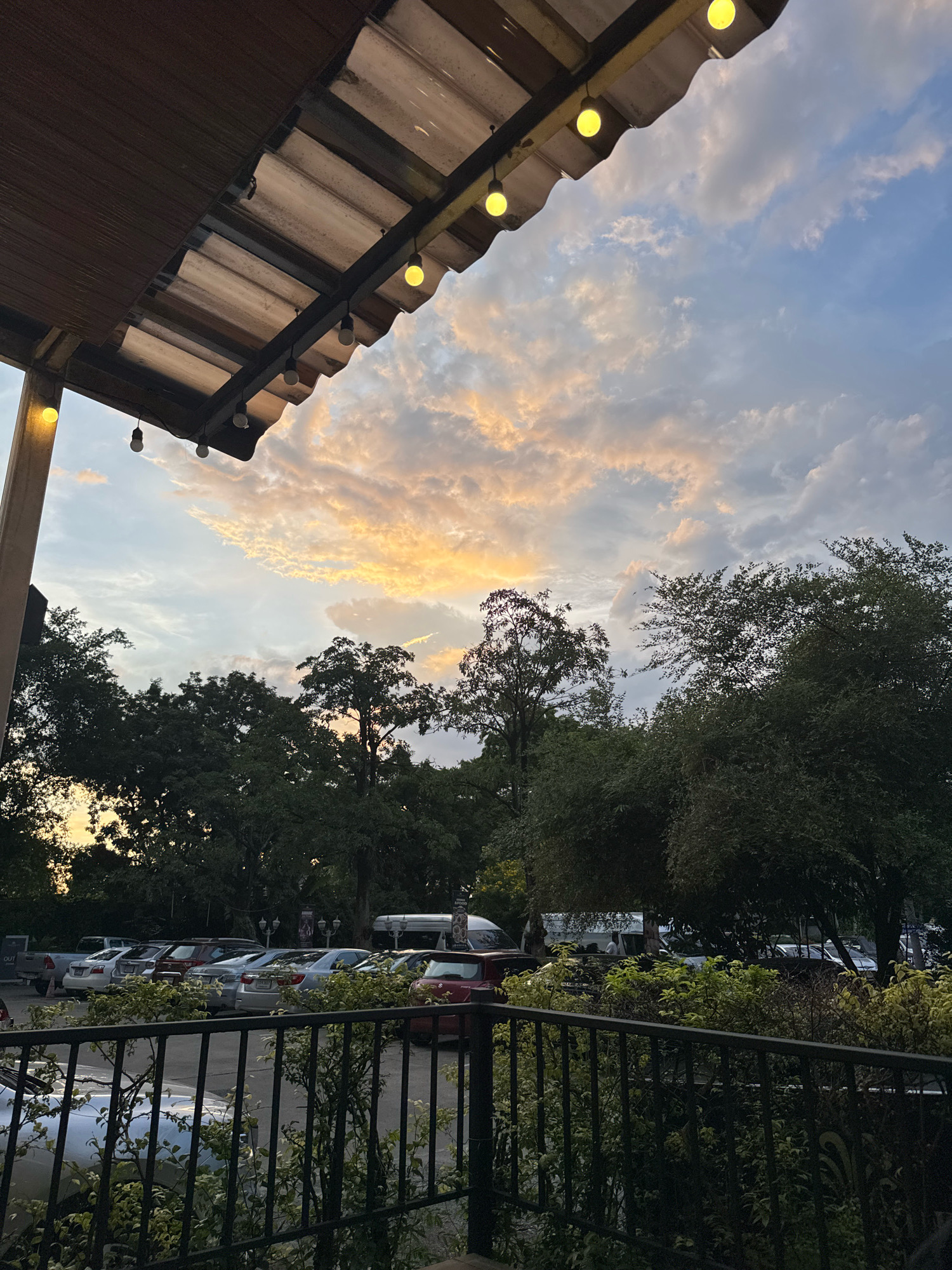 The image depicts an outdoor scene, likely a dining or gathering area, under a covered roof with exposed light bulbs. In the background, a parking lot filled with various cars is visible, bordered by lush green trees. Beyond the trees, the sky displays a sunset with clouds illuminated in hues of orange and yellow, transitioning to blue. The foreground features a black metal fence and some vegetation. The setting appears to be a restaurant or a similar establishment in Bangkok, Thailand, given the style of the roof and the tropical foliage. No individuals are clearly discernible.