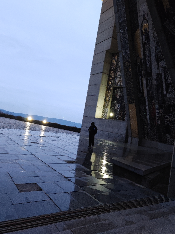 The image depicts an outdoor scene at the Monument to 1300 Years of Bulgaria in Shumen, Bulgaria, observed at twilight or dawn. A large, angular concrete monument, partially illuminated by spotlights, dominates the right side of the frame. Its surfaces feature dark, intricate mosaic murals depicting historical figures and scenes.
A solitary individual, dressed in dark attire, stands facing away from the camera near the base of the monument, positioned between two illuminated sections. The ground is paved with dark, rectangular tiles and appears wet, exhibiting significant reflections of the monument's lights and the individual's silhouette. Some tiles show signs of damage or absence. In the immediate foreground, a series of steps is visible.
Beyond the paved area and the monument, a distant landscape of low-lying hills or mountains is visible under a pale, overcast sky. Two distinct bright lights, likely artificial, are present on the distant horizon. The overall lighting is low, with artificial illumination contributing to contrast and reflections on the wet surfaces. No other individuals or discernible activities are present.