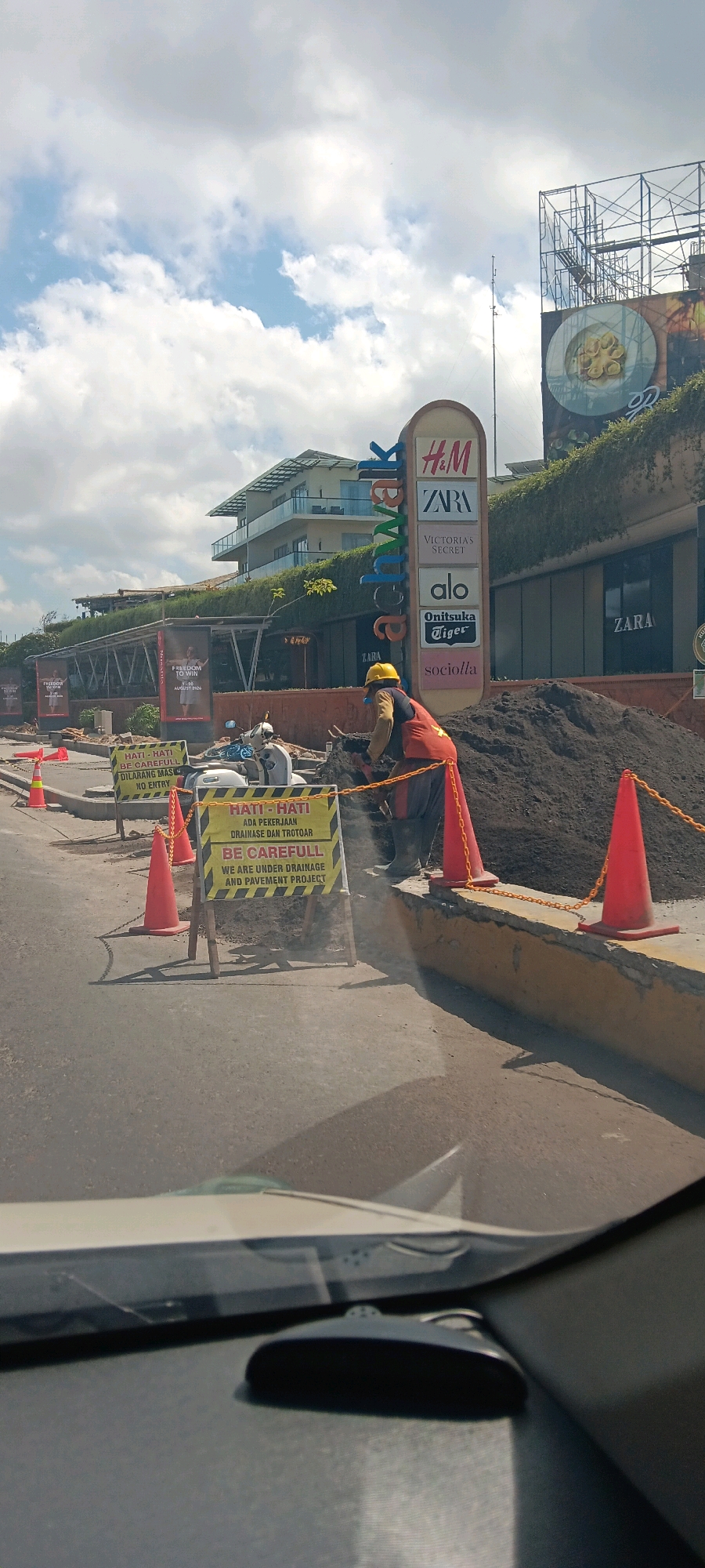 The image shows a construction site in Kuta, Bali, Indonesia, with a prominent "Beachwalk" sign displaying various brands like H&M, Zara, Victoria's Secret, Alo, Onitsuka Tiger, and Sociolla. A worker in a yellow hard hat and an orange vest is operating a cutting tool, likely for pavement, near a large pile of dirt. Orange traffic cones and chains delineate the construction area. Several warning signs in both Indonesian and English are visible, cautioning passersby about drainage and pavement projects and advising "No Entry". In the background, a multi-story building with a modern architectural design and lush greenery is visible, suggesting it's part of a shopping complex. The sky is partly cloudy, indicating daytime. A scooter is parked near the construction area, and the foreground shows a car's dashboard and windshield.
