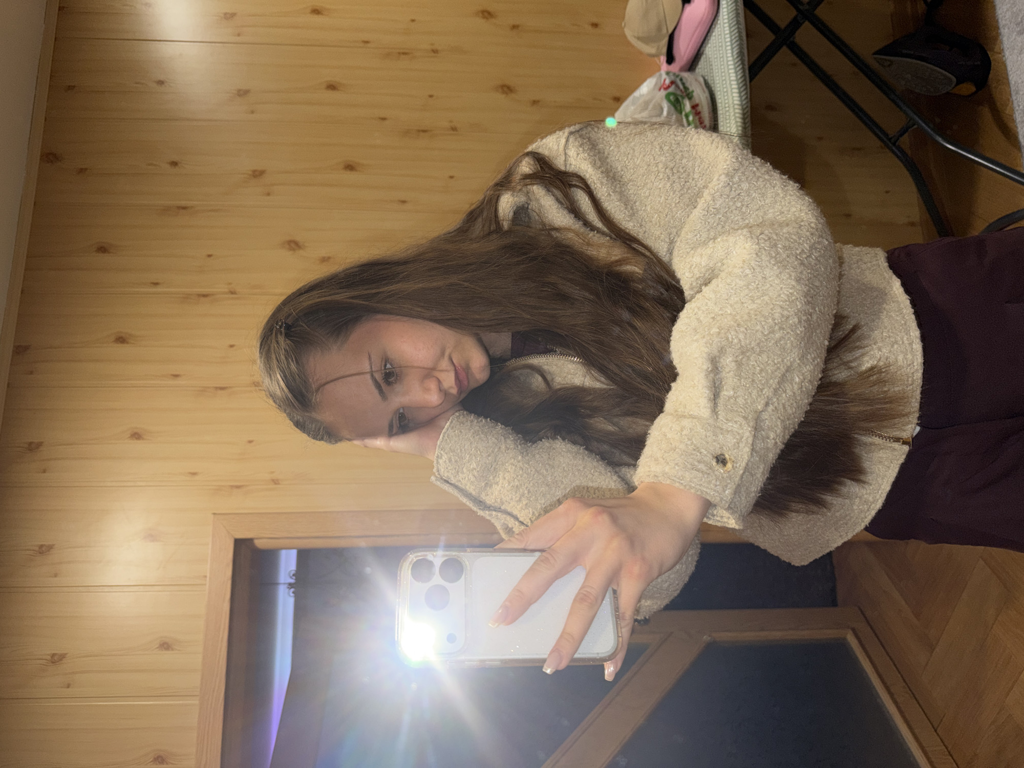 A young woman with long brown hair is holding a white smartphone with its flash on, taking a selfie. She is wearing a light-colored, textured jacket and a dark burgundy top. The background consists of wood paneling on the left, a doorway in the lower-left corner, and wooden flooring on the right. Above her, a small table is visible with items including a pink hat and a grocery bag on it, as well as a black stool. There is no indication that the photo was taken in Buhovo, Bulgaria.