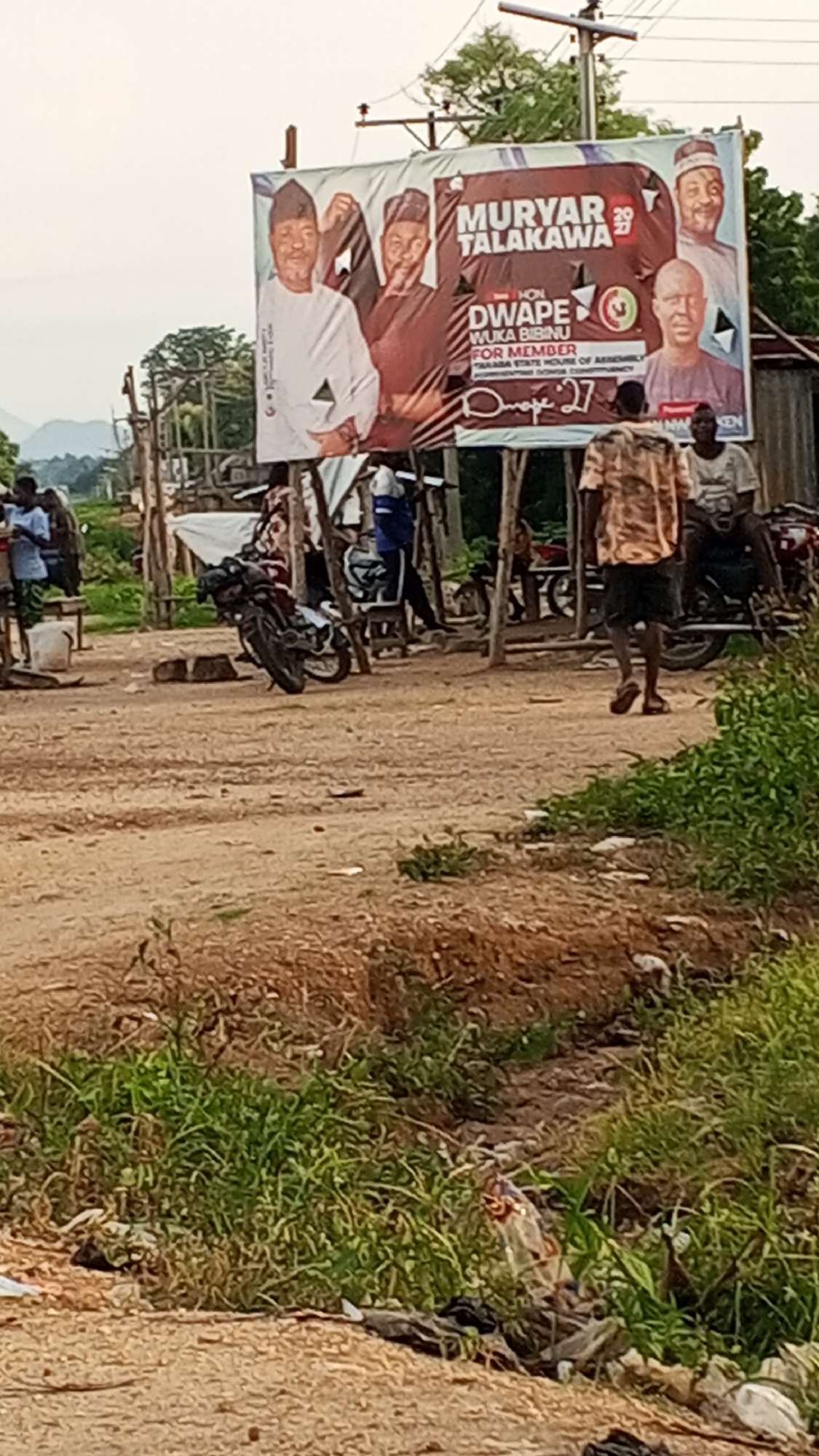 In Nyita, Nigeria, a large billboard displays a political campaign for "MURYAR TALAKAWA" and "DWAPE BIBINU FOR MEMBER". Several individuals are visible in the scene. One person, wearing a patterned short-sleeved shirt and shorts, walks away from the viewer towards the right. Another person, seated to the right of the billboard, wears a light-colored t-shirt and dark shorts. In the background, several people are gathered around makeshift stalls, some with motorcycles present. The environment appears to be a dusty, open area with vegetation and scattered debris, suggesting a rural or semi-urban setting. The billboard is supported by wooden poles. The overall impression is that of a public gathering or marketplace where political messaging is prominent.