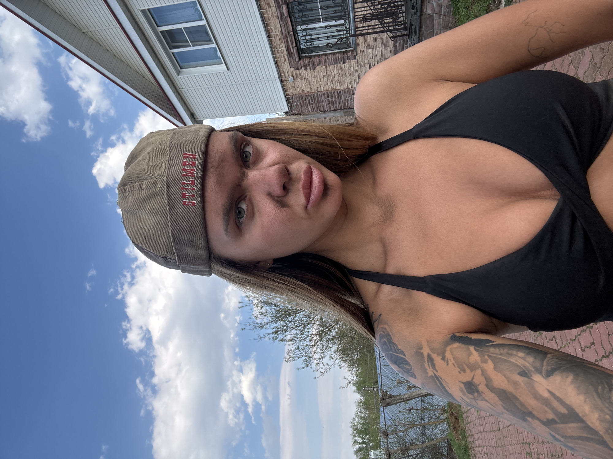 A woman in a black top and a cap with the word "STYLIST" is posing for a selfie in Bol'shoye Kozino, Russia. The background includes a house, a cloudy sky, and greenery. She has tattoos on her arms. The area is paved.