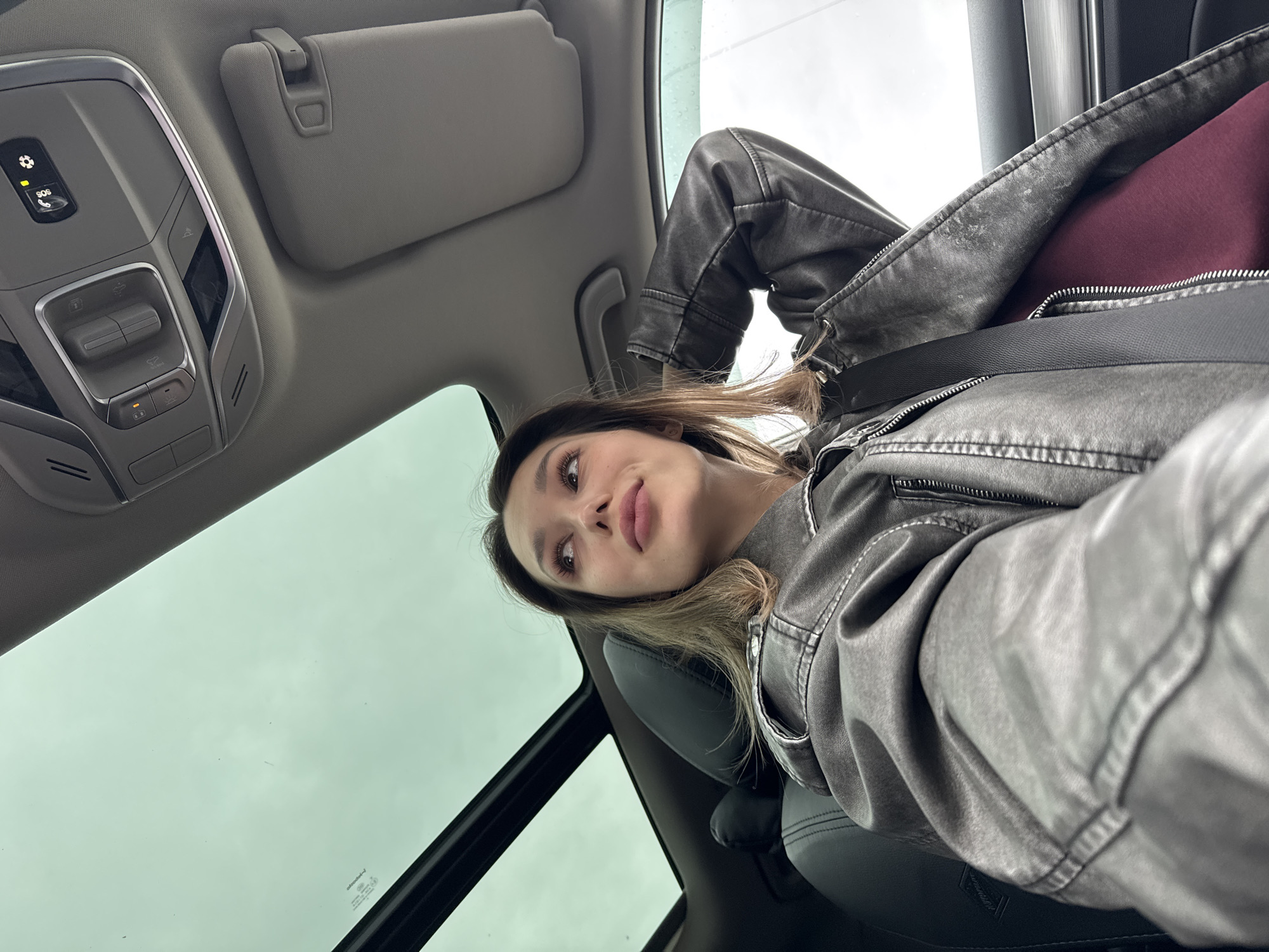 A woman is sitting in a car, wearing a grey leather jacket and a maroon shirt, and is secured with a seatbelt. The car has a sunroof, through which a cloudy sky is visible. The car's interior features overhead controls and a sun visor. The location is undetermined.