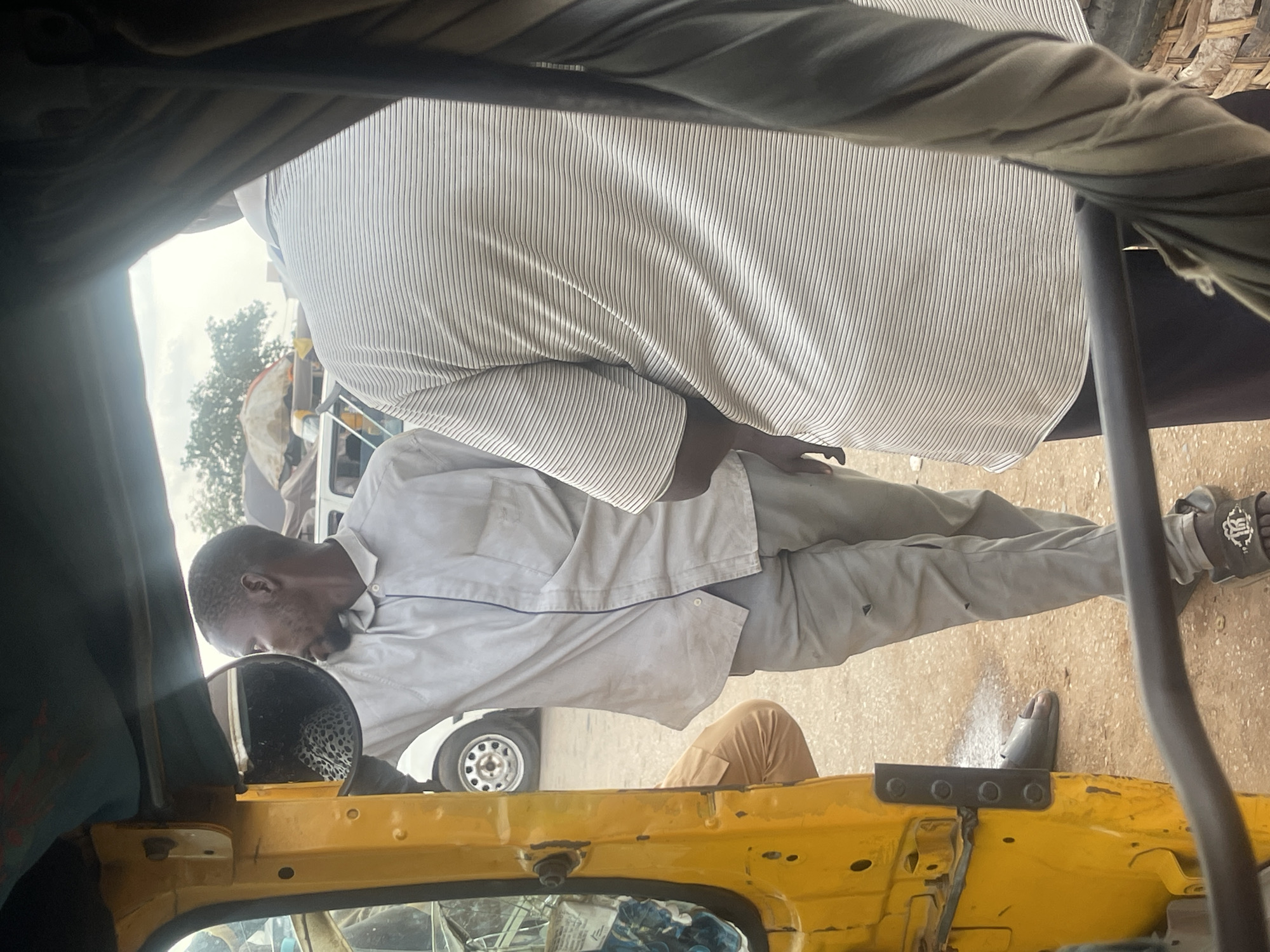 The image shows two individuals in what appears to be an outdoor setting, possibly a village in Nigeria. The foreground is dominated by the yellow interior of a vehicle, likely a tuk-tuk or similar motorized rickshaw, with parts of its dashboard and door visible.
A man in a light-colored, possibly linen, shirt and trousers is leaning towards a side mirror. He has short dark hair and a beard. His posture suggests he is either inspecting the mirror or engaged in a task related to the vehicle. His left hand is near the mirror, and his right arm is bent. He is wearing sandals.
In the background, another person is partially visible, wearing a horizontally striped garment. This individual is seated or standing behind the first man, closer to the vehicle's roof. The background shows some greenery and trees, indicating an outdoor environment, and a white vehicle is parked behind the man in the foreground. The lighting suggests it is daytime, with the sun casting rays, indicated by the glare on the mirror and a bright sky. The overall impression is that of a casual, possibly work-related, scene in a rural or semi-rural Nigerian location. No text is visible in the image.