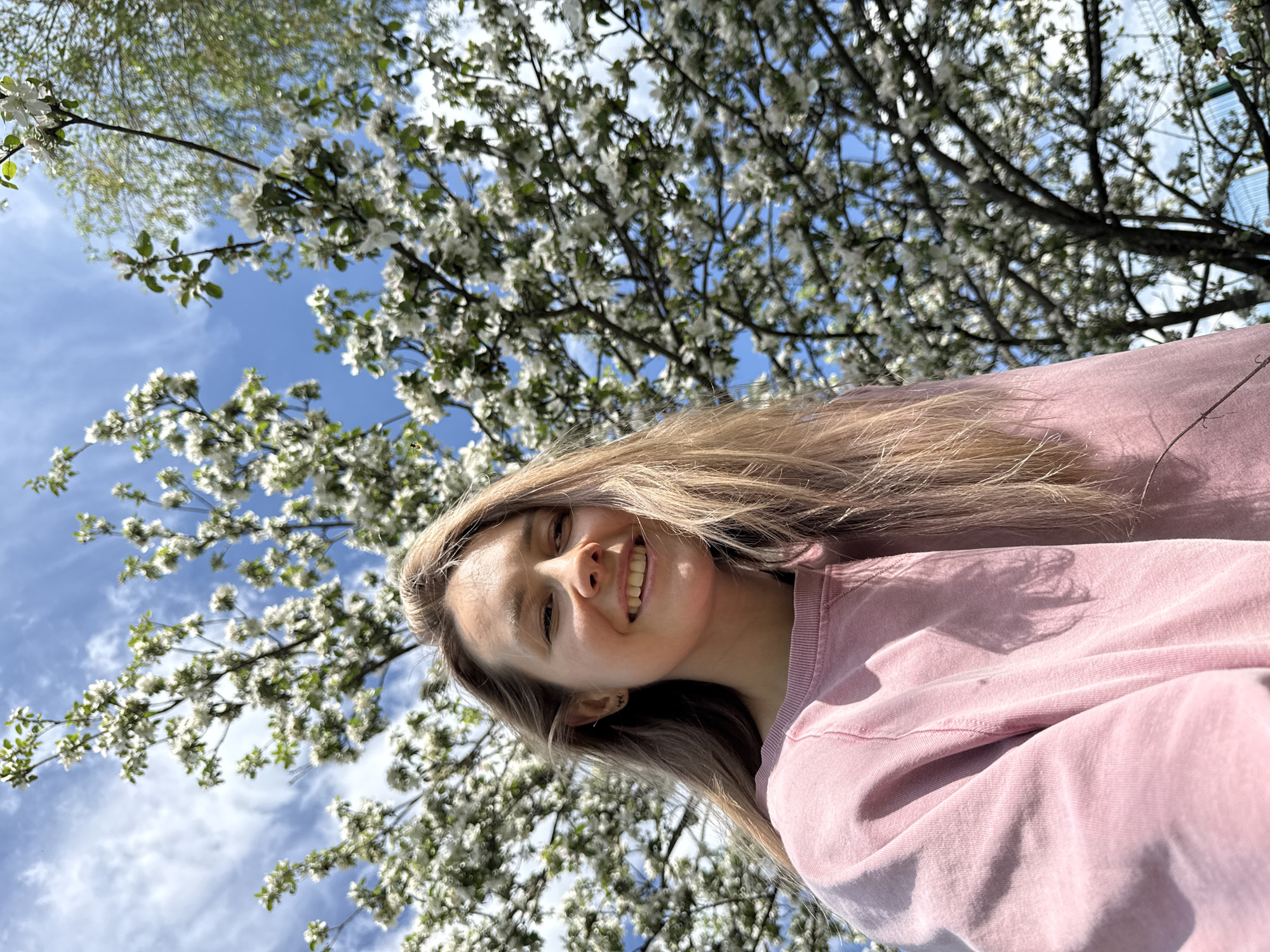 A young woman smiles in a selfie. She has long, light brown hair and is wearing a pink sweatshirt. Behind her is a tree with white blossoms, and blue sky is visible. The location cannot be identified as Noviy, Russia from the image alone.