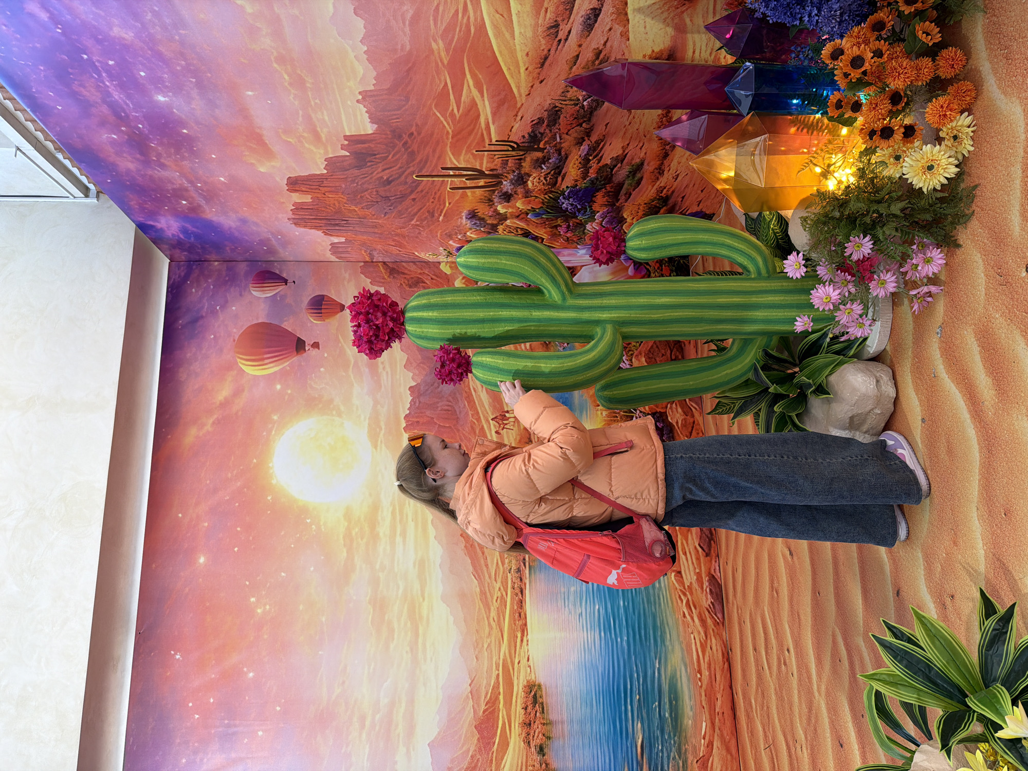 The image shows a person standing in front of a desert-themed installation. The installation has a backdrop that features a sunset over a desert landscape with hot air balloons. The ceiling above depicts a star-filled sky. There is a large inflatable cactus with pink flowers attached to it. Other elements in the installation include colorful crystal shapes, and arrangements of various flowers. The person is wearing an orange puffer jacket, blue jeans, and sneakers. They have a red backpack slung over their shoulder. Based on the presence of Berezniki signage in related search results and the consistent desert themed installation, this photo was likely taken in the city of Berezniki, Russia.