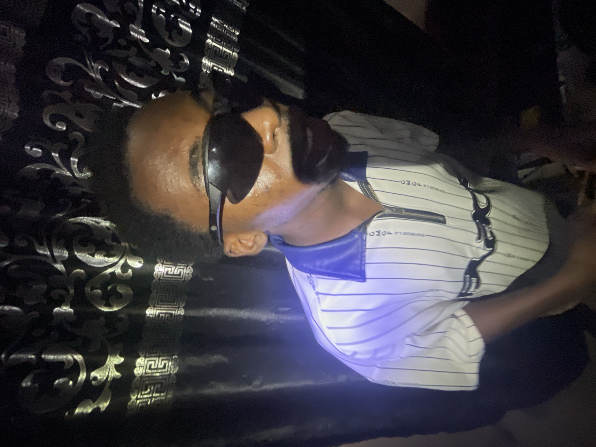 A person wearing sunglasses and a striped shirt is seated against a dark, ornate backdrop. The location is difficult to determine definitively but if in Lokoja, Nigeria, it's likely an indoor location. The person appears to be posing for the photo, and the environment seems dimly lit. More context is needed for a more precise description.