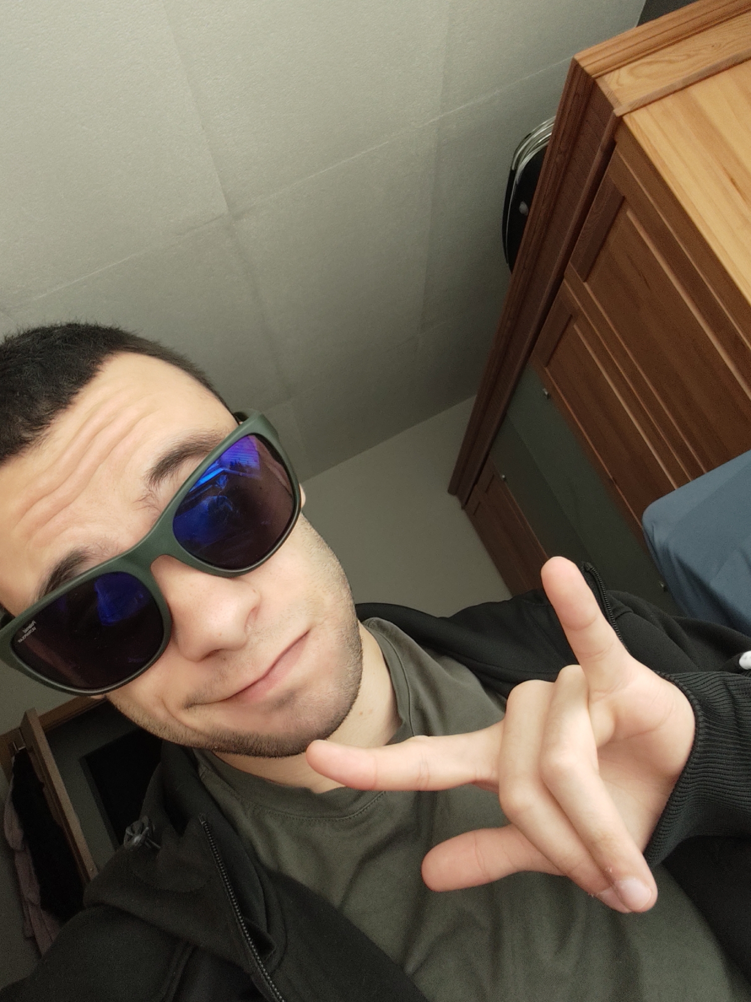 A young adult male is captured in a close-up, eye-level selfie, taken indoors in what appears to be a room in Sofia, Bulgaria. He is looking towards the camera with a slight head tilt. He wears dark-framed, reflective sunglasses with blue-purple lenses, which obscure his eyes but reveal a subtle smirk around his mouth. The left inner arm of the sunglasses shows visible text, including "UV Protection" and "RADIAN". He has short dark hair and light stubble.
He is dressed in a casual olive green t-shirt underneath a black zip-up hoodie. His right hand is raised, performing a common hand gesture where the index and pinky fingers are extended, and the middle and ring fingers are curled.
The background features a plain, off-white ceiling and walls. To the right, a large, light-brown wooden wardrobe or cabinet with paneled doors is visible. Partially hidden behind the wardrobe is a dark object, possibly a piece of luggage or a bag. In the lower right background, a dark blue or grey fabric, possibly a duvet or blanket on a bed, can be seen. The lighting in the room is bright and even, indicating a daytime setting with adequate indoor illumination. The overall impression is casual and confident.