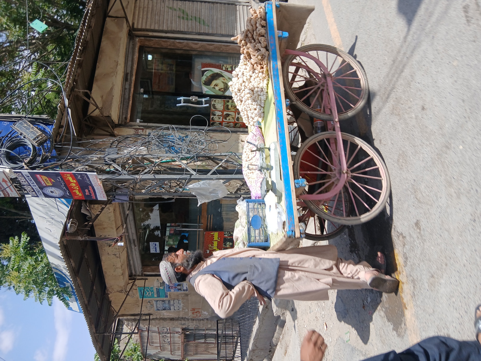 The image depicts a man standing on a street in Quetta City Tehsil, Pakistan, beside a wheelbarrow loaded with produce. The wheelbarrow has pink wheels and is piled high with what appears to be ginger and other items. The man is wearing traditional Pakistani clothing, including a vest and cap. Behind the man and the wheelbarrow is a building with a cluttered facade, including a complex web of electrical wires and various signs and posters. A poster in the window shows images of men's haircuts, which may indicate a barbershop. The road is asphalt.