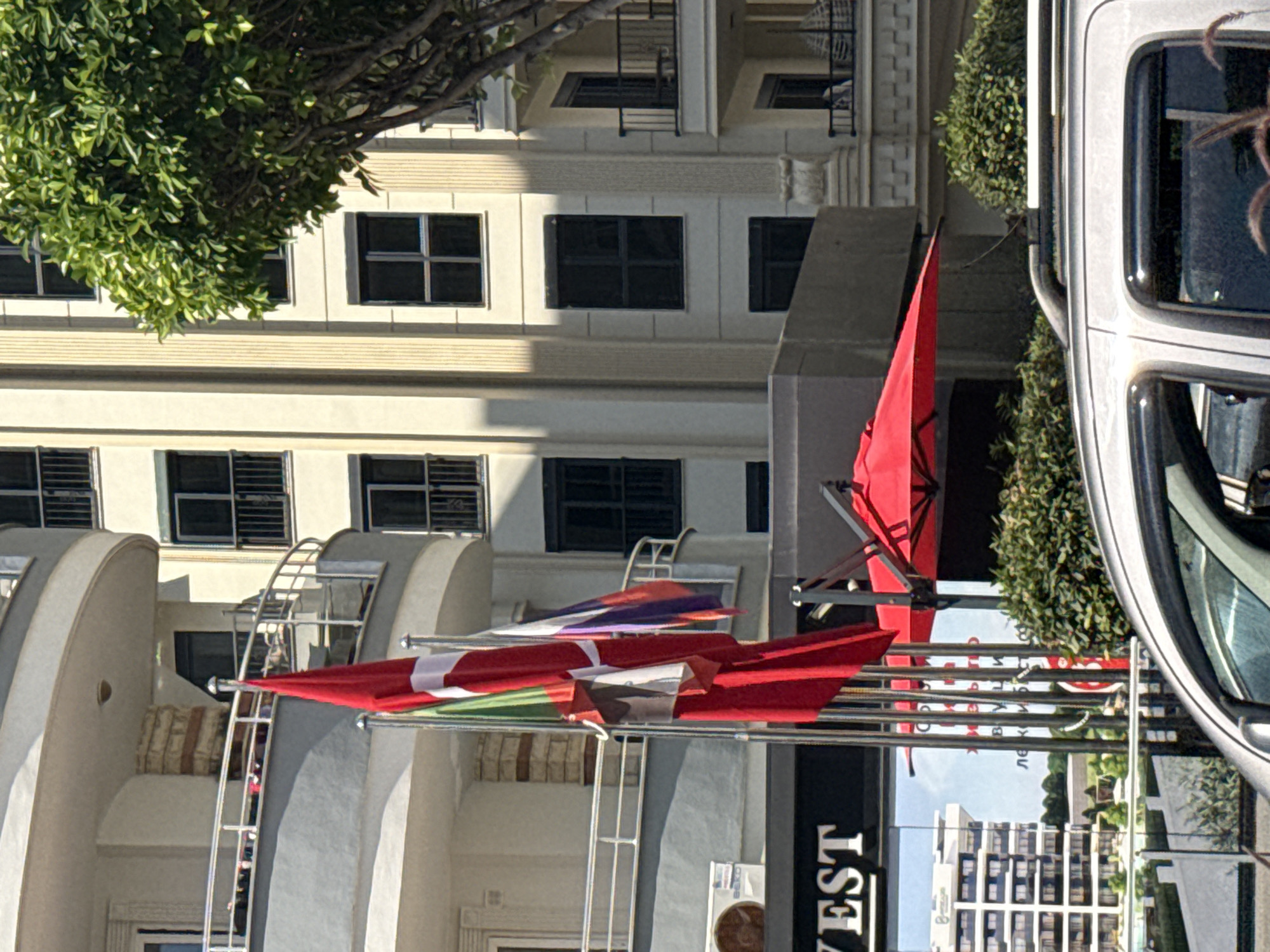 The image shows a street scene in an urban area, possibly in Unknown, Unknown, featuring a building with multiple windows and architectural details like rounded facades and ladders. Several flags, predominantly red and one with a white cross, are on display. A white vehicle is partially visible on the right side of the frame, and a tree with dense foliage is in the upper left corner. Additionally, a sign with the word "VEST" is present, along with other buildings across the street in the background.